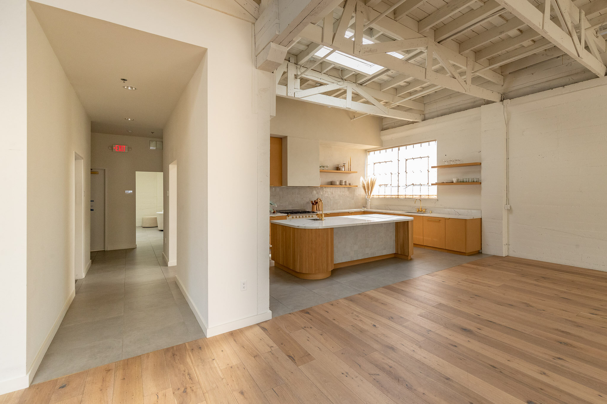 Open-concept kitchen with a wooden island, open shelves, large window, exposed ceiling beams, hardwood flooring, and a hallway leading to other rooms.