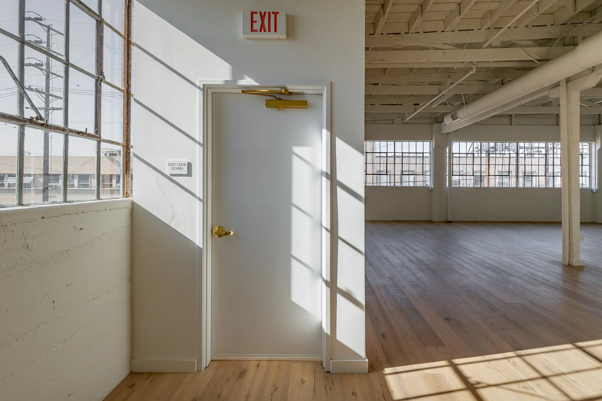 Empty room with large windows, an exit door with a gold handle, a small sign indicating 'Exit Stair Down,' and sunlight casting shadows on the white walls and wooden floor.