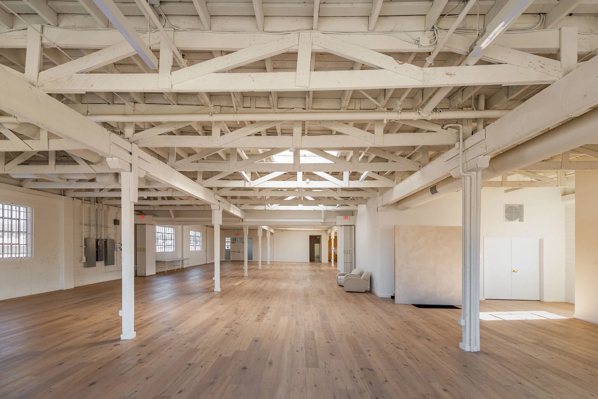 Spacious, empty indoor room with high white ceiling exposed beams, large windows letting in natural light, light wood flooring, minimal furniture, and white walls.
