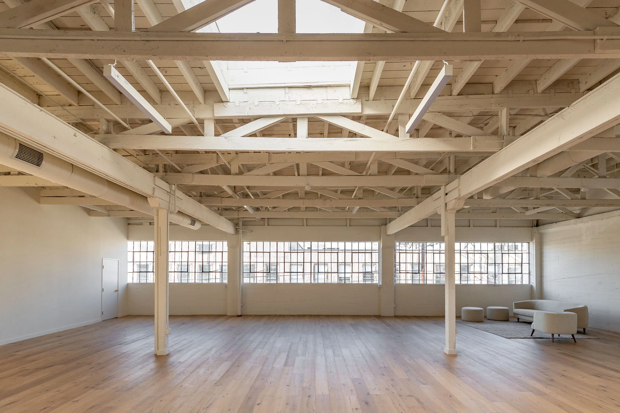 Spacious empty room with large windows, wooden floor, exposed white ceiling beams, and modern white furniture in the corner.