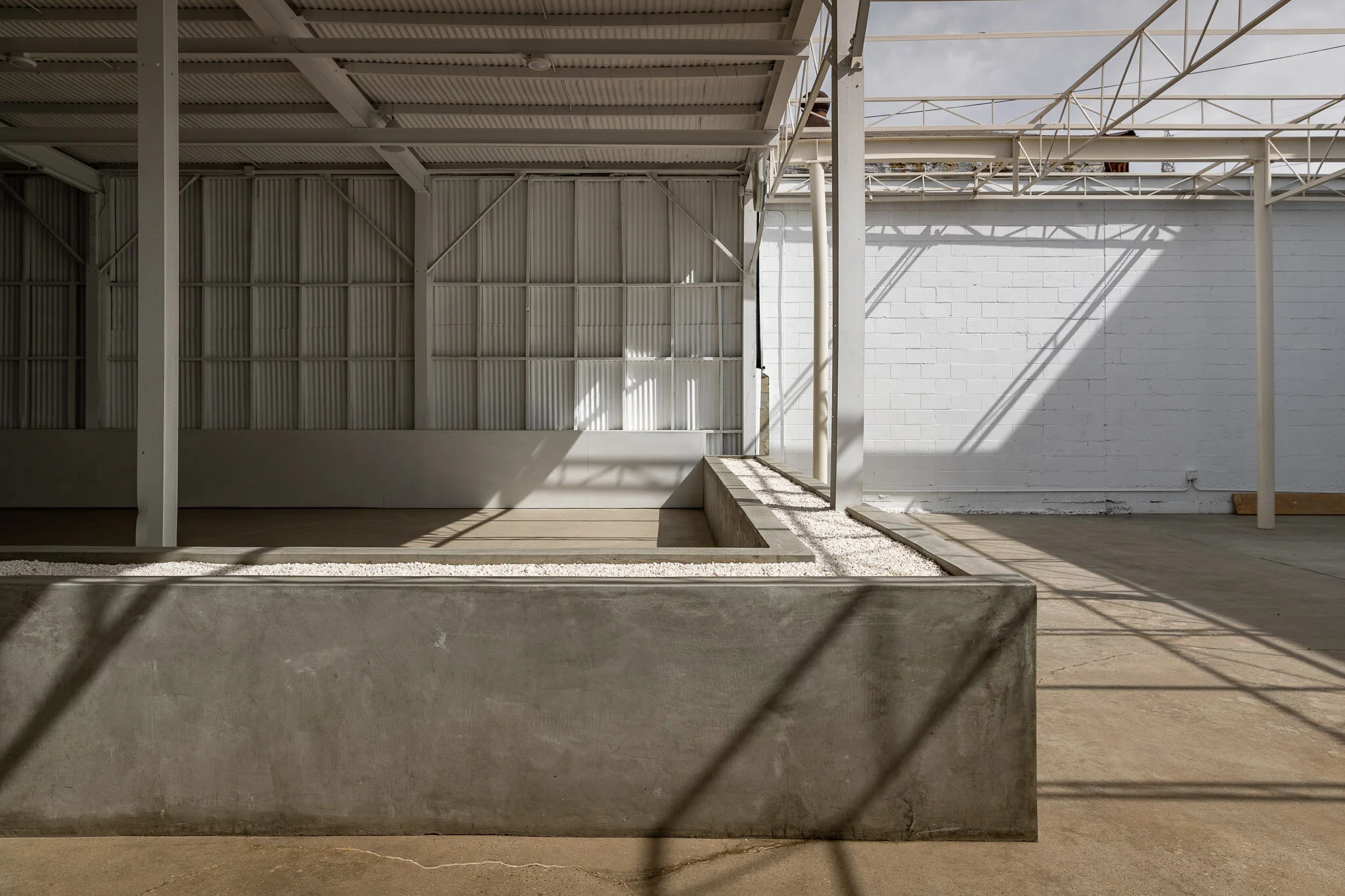 Empty indoor space with white walls, metal roof, shadows from a partial outdoor structure, and a small elevated garden bed with a gravel border.