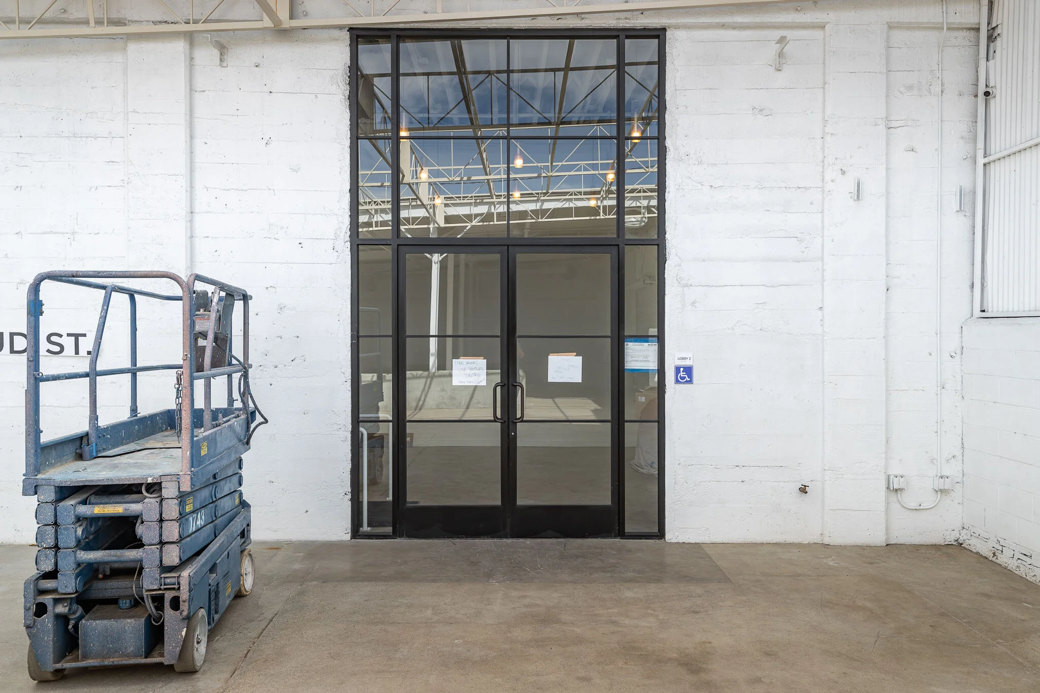 Industrial warehouse entrance with large glass doorway, white brick walls, a blue handicap parking sign, and a blue scissor lift parked to the left.
