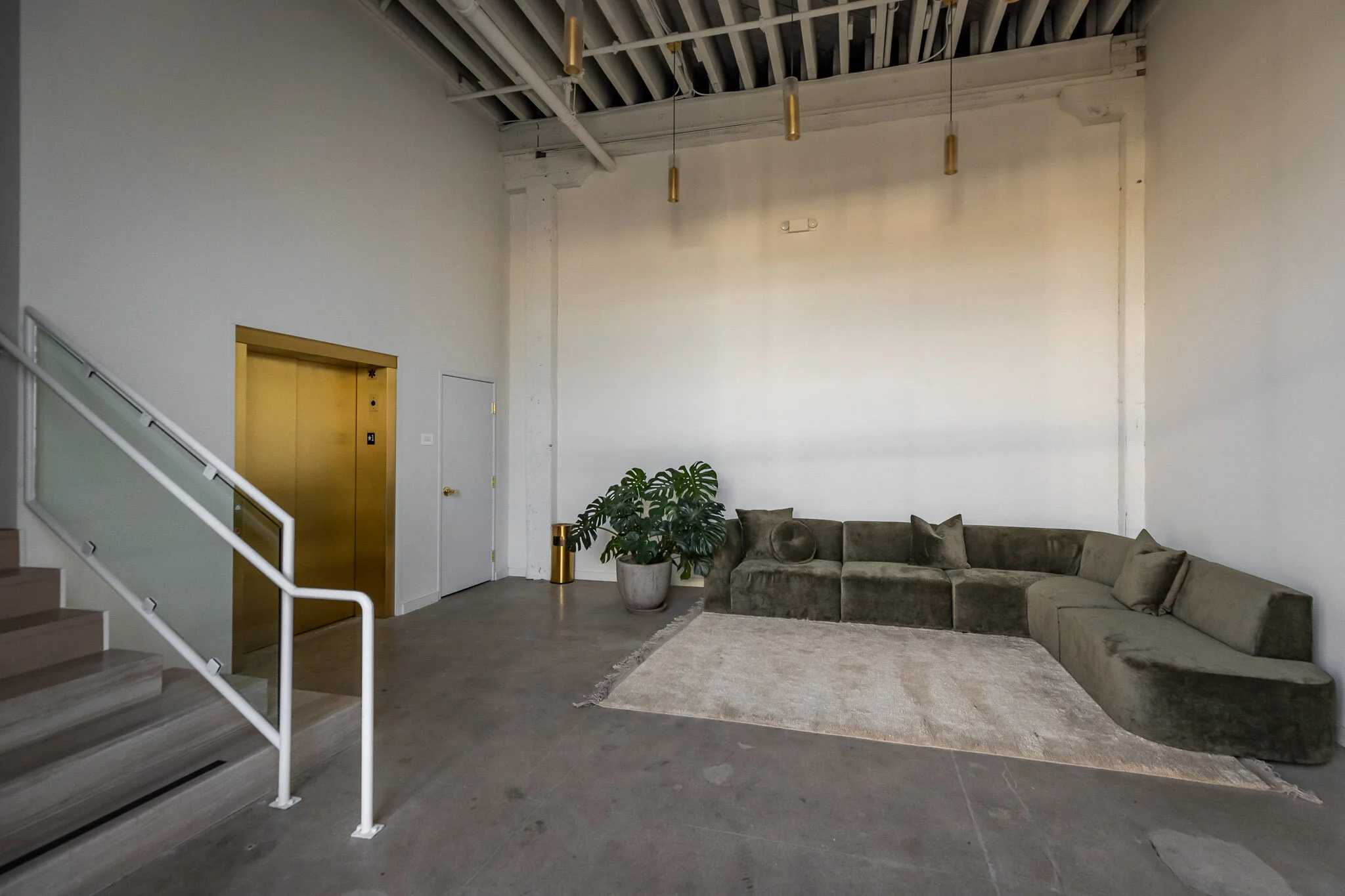 Modern lounge area with a beige sectional sofa, green indoor plant in a gray pot, a gold-colored trash bin, and a light-colored rug on an industrial-style concrete floor. Gold pendant lights hang from the ceiling, with an elevator and stairs nearby.