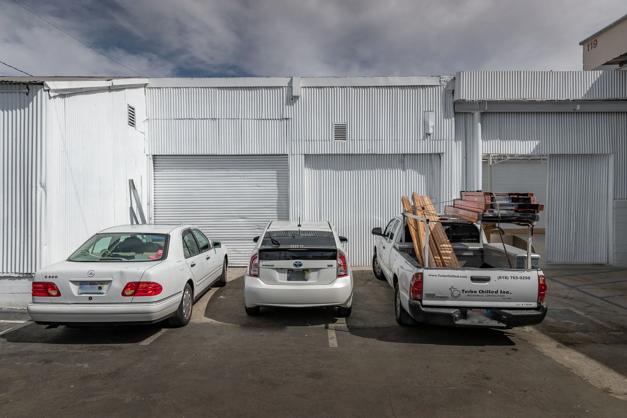 Three vehicles parked in front of a white industrial building with metal siding and a large garage door, with construction lumber in the truck bed on the right.
