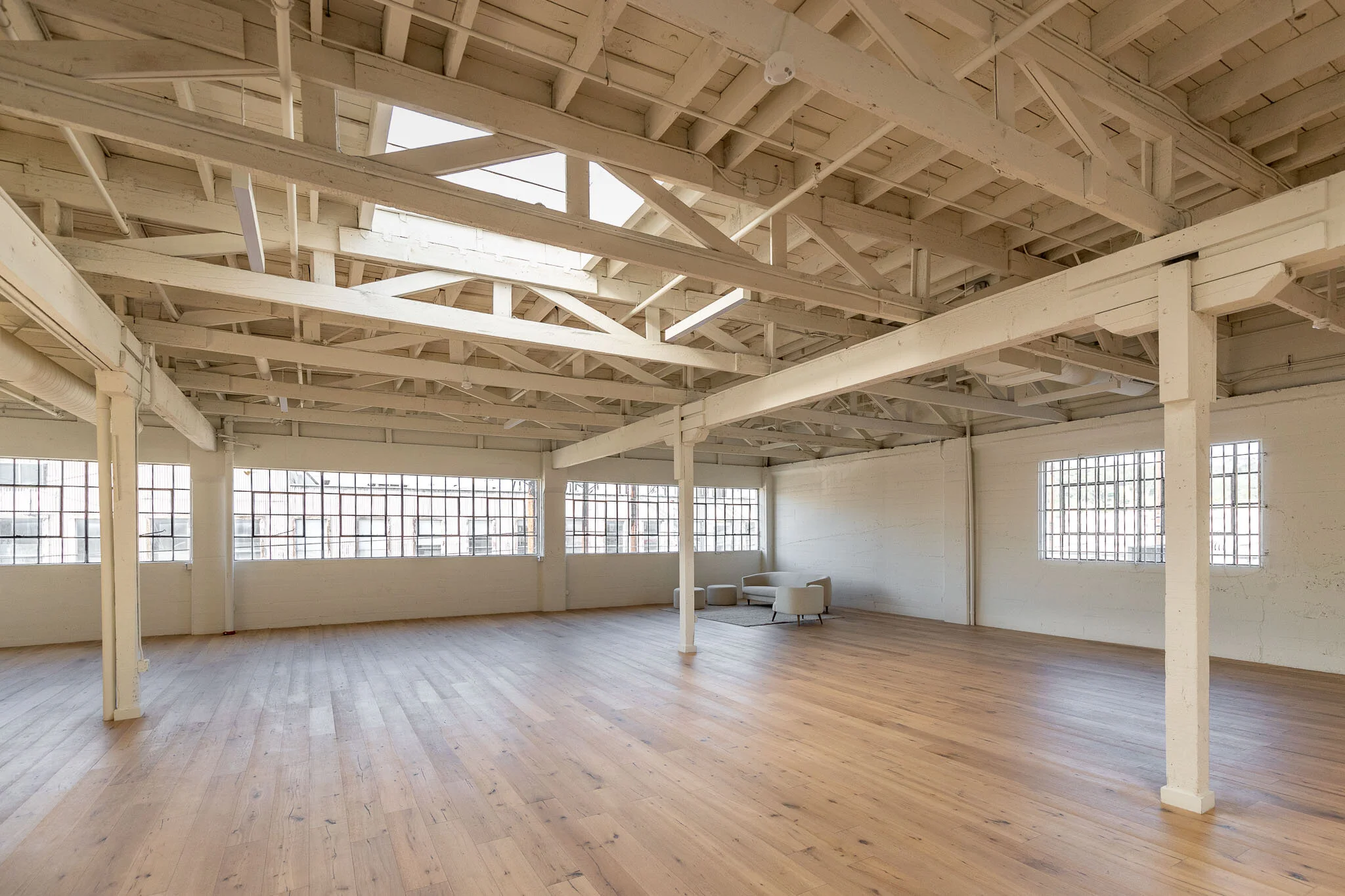 Spacious indoor area with white-painted exposed beams and large windows, hardwood flooring, minimal furniture including a white sofa, ottomans, and a small table near the wall.