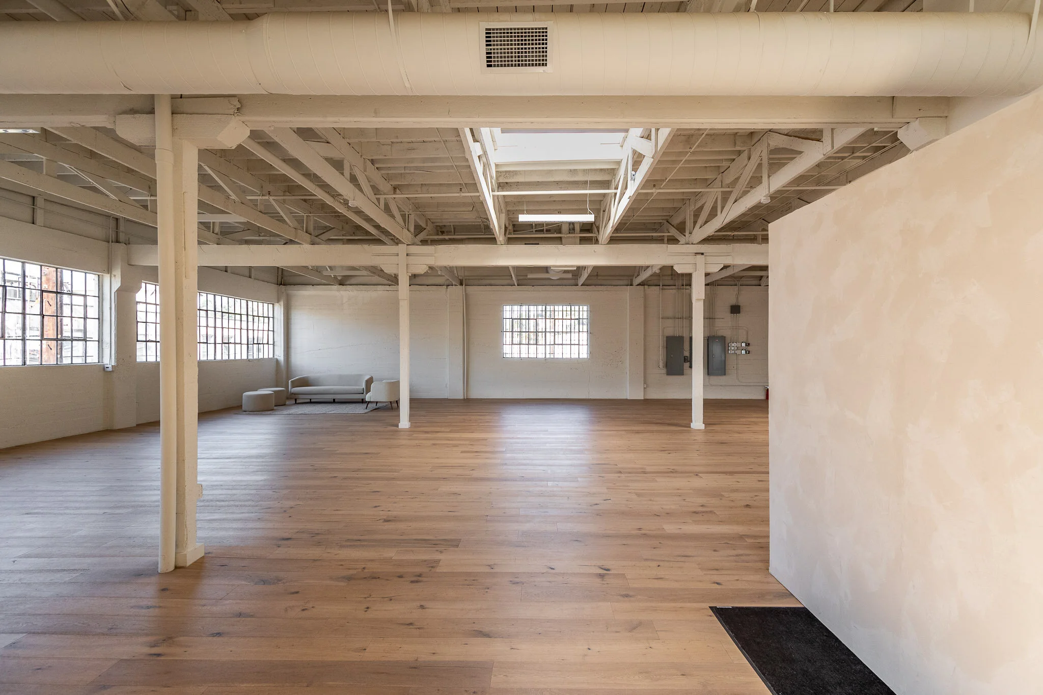 Empty industrial-style room with white walls, large windows, wooden floors, and a white couch with a matching ottoman in the corner.