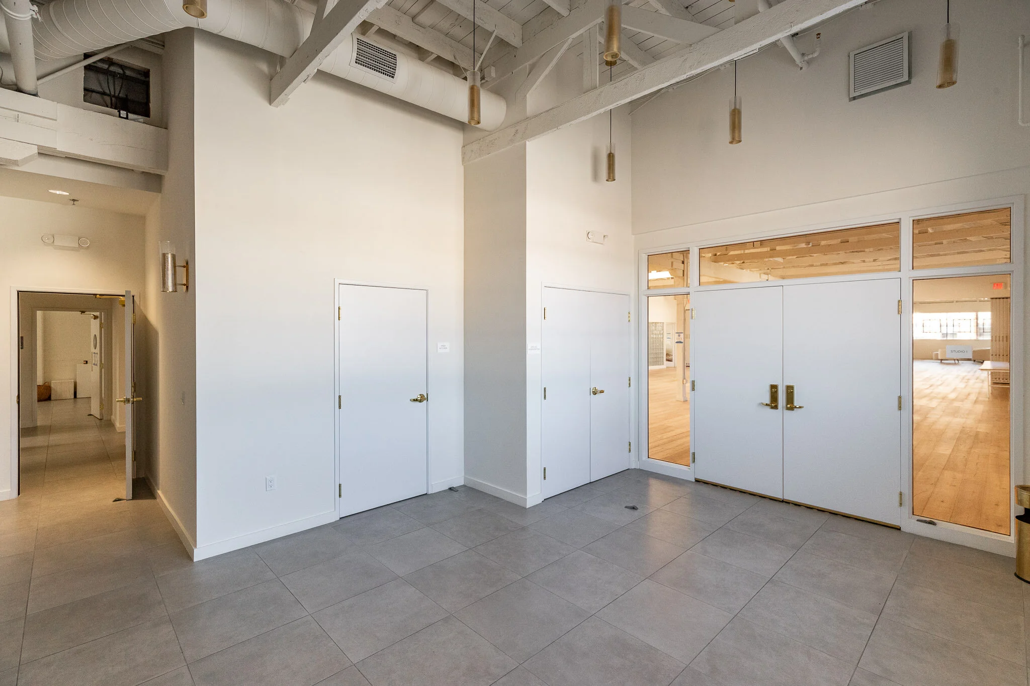 Empty interior room with white walls, tiled grey floor, multiple white doors, large windows, and exposed ceiling beams.