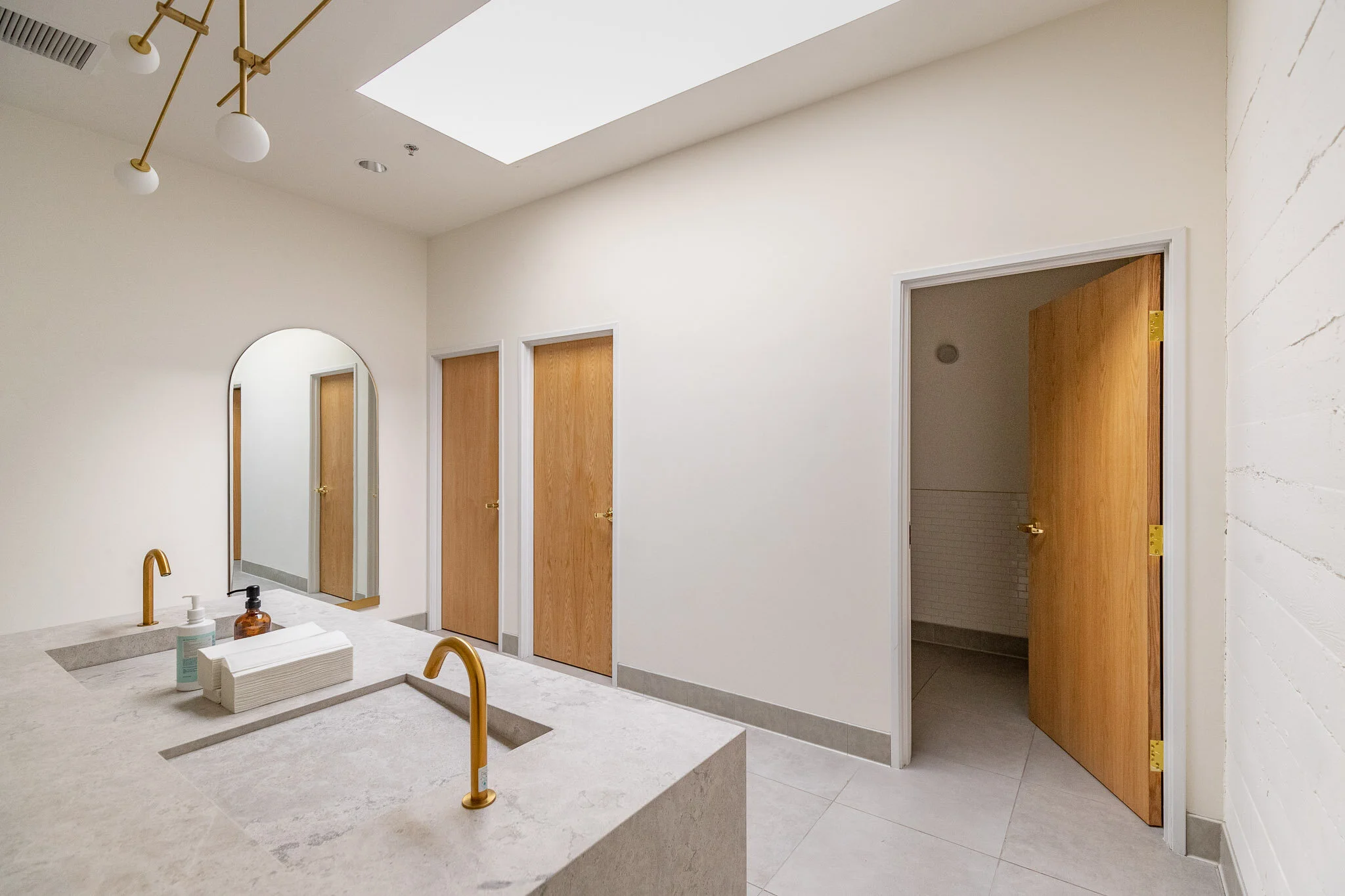 Modern bathroom with two sinks, gold faucets, hand soap, paper towels, a large mirror, ceiling lighting, wooden doors for stalls or storage, and a skylight bringing in natural light.
