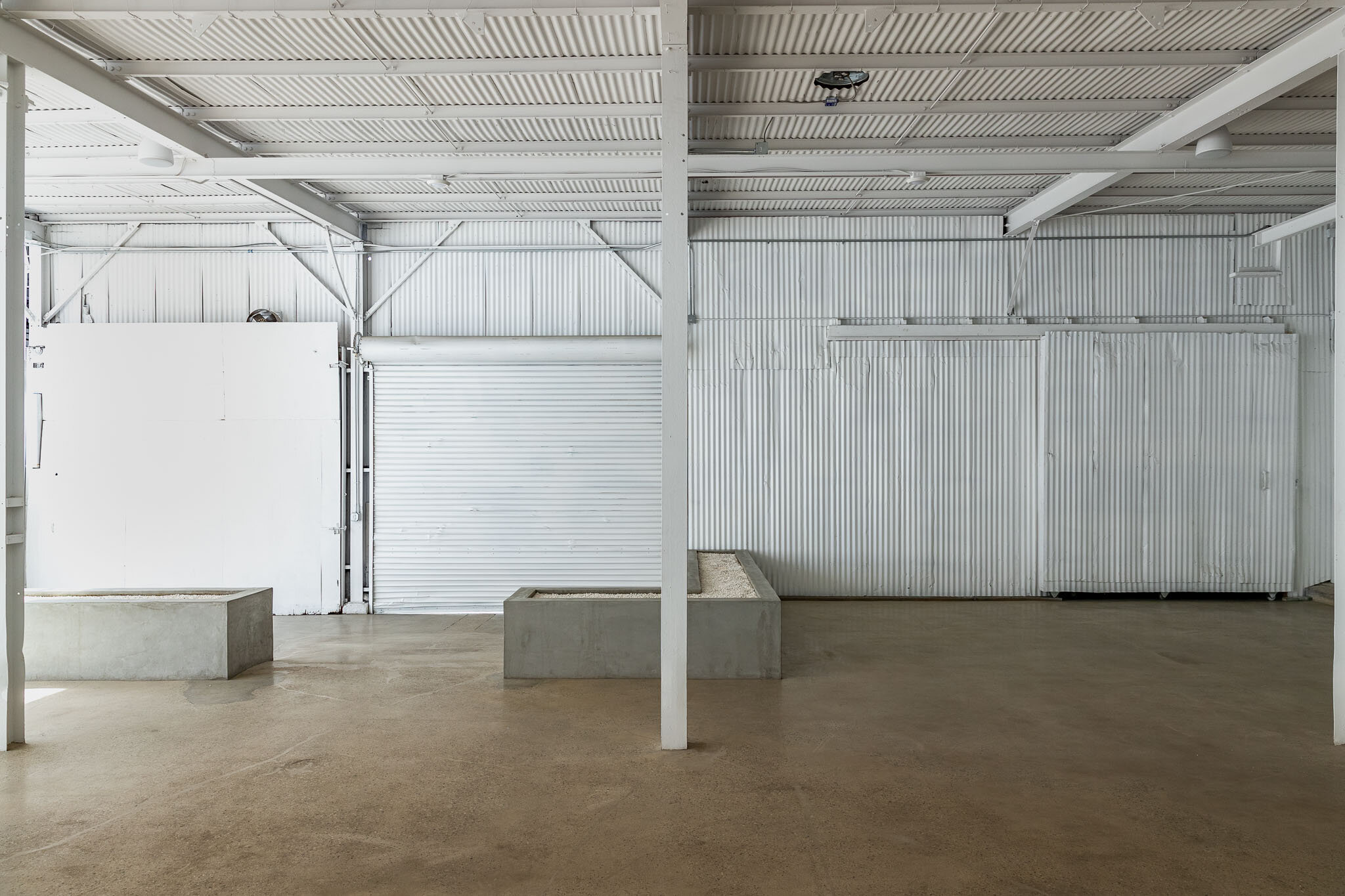 Empty industrial warehouse with metal walls, concrete planters, and a beige concrete floor.