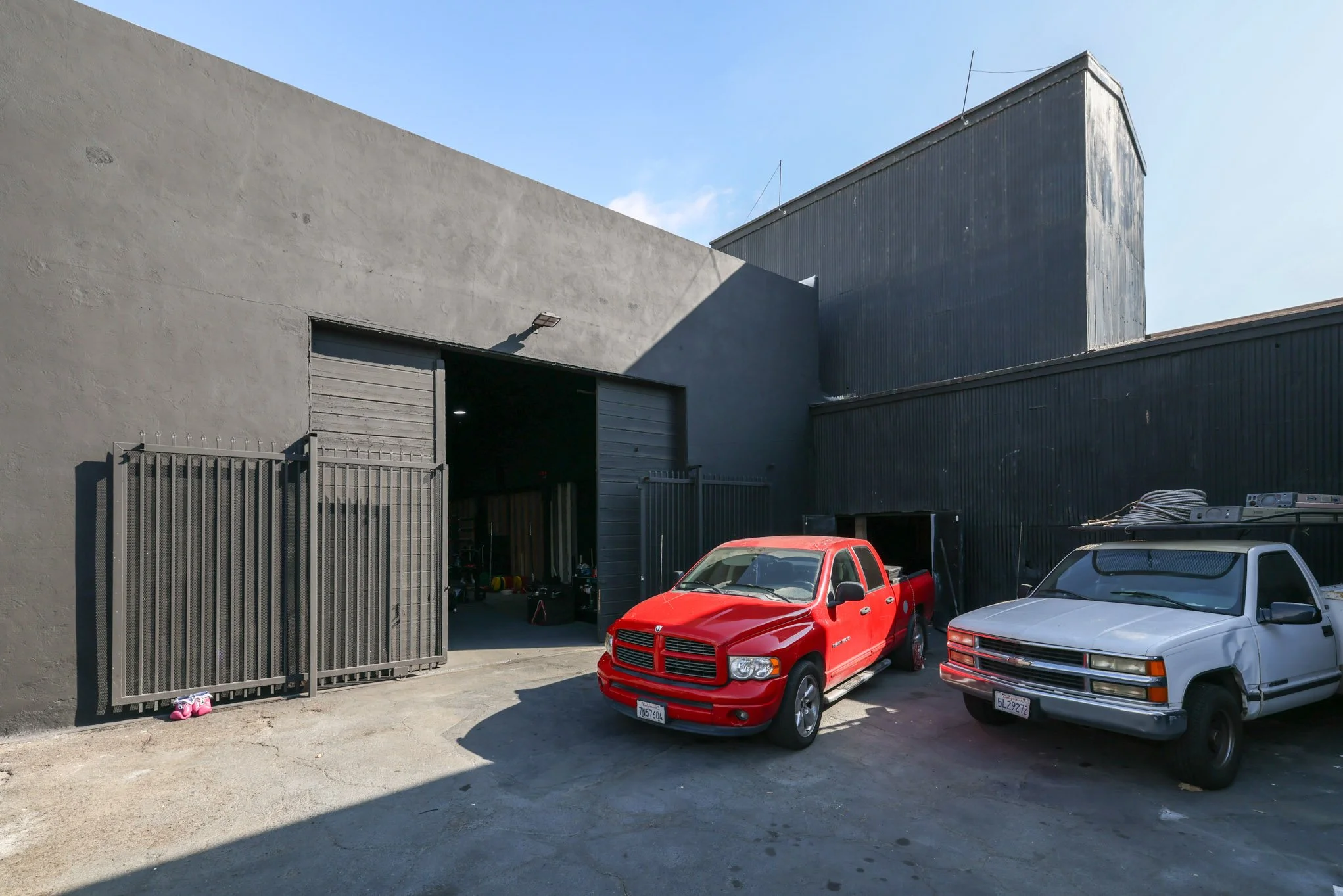 A red pickup truck and a white van parked outside a gray industrial building with open garage doors on a sunny day.