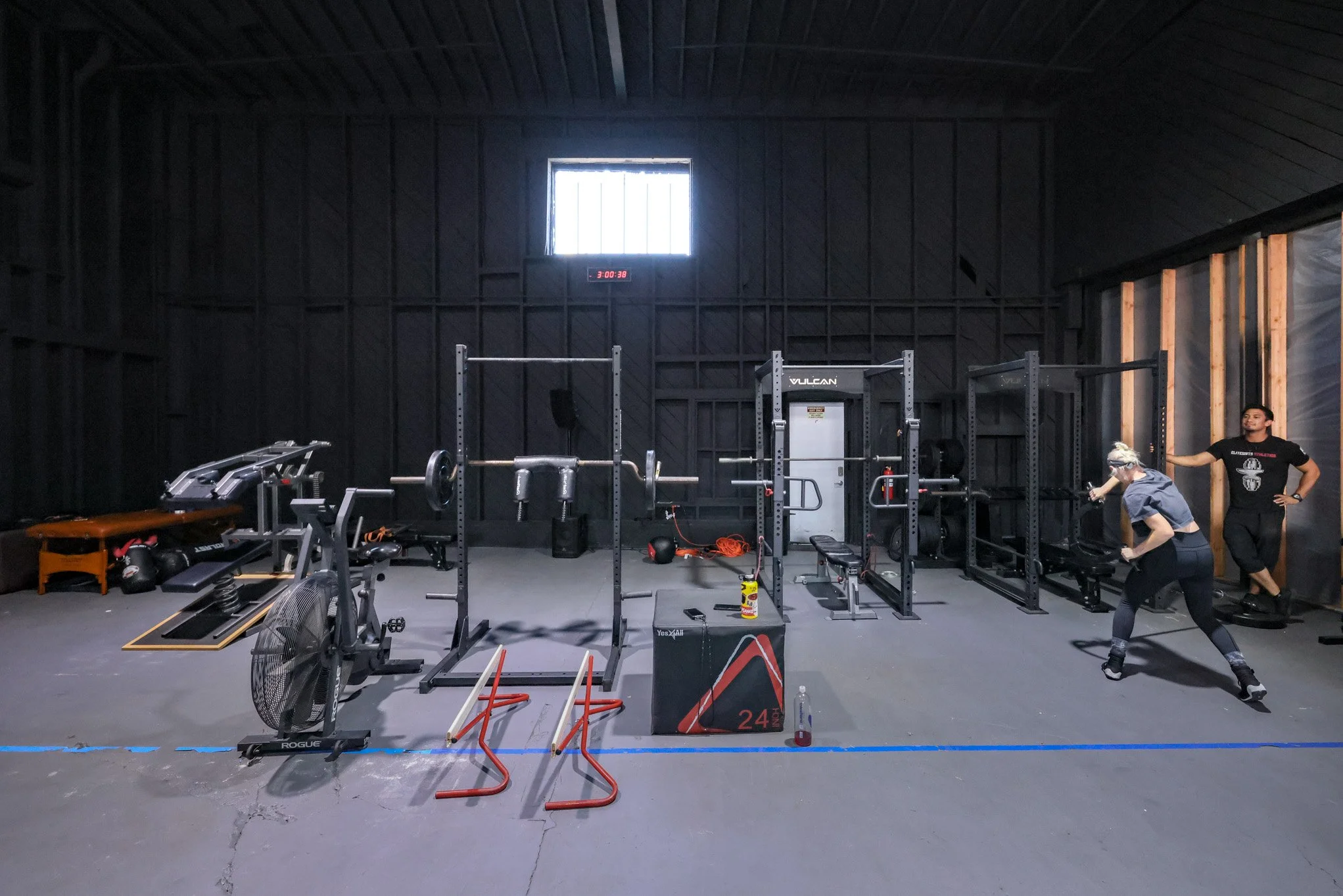 Indoor gym with black walls and various exercise equipment including weight racks, a bench, a fan, and resistance bands. Two people are stretching in the right corner, with one man standing and the woman in a stretch pose.