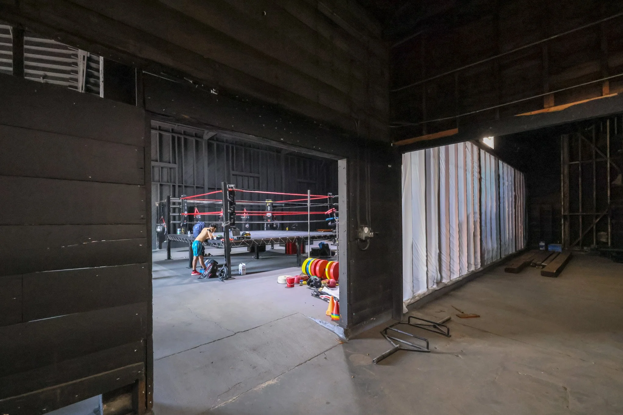 Inside a boxing gym with a boxing ring, punching bags, and various training equipment. Two people are preparing for training near the ring.