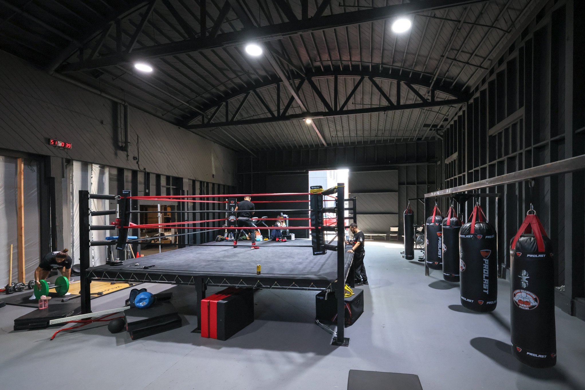 An indoor boxing gym with a boxing ring in the center, several black punching bags hanging on the right wall, and people working out.
