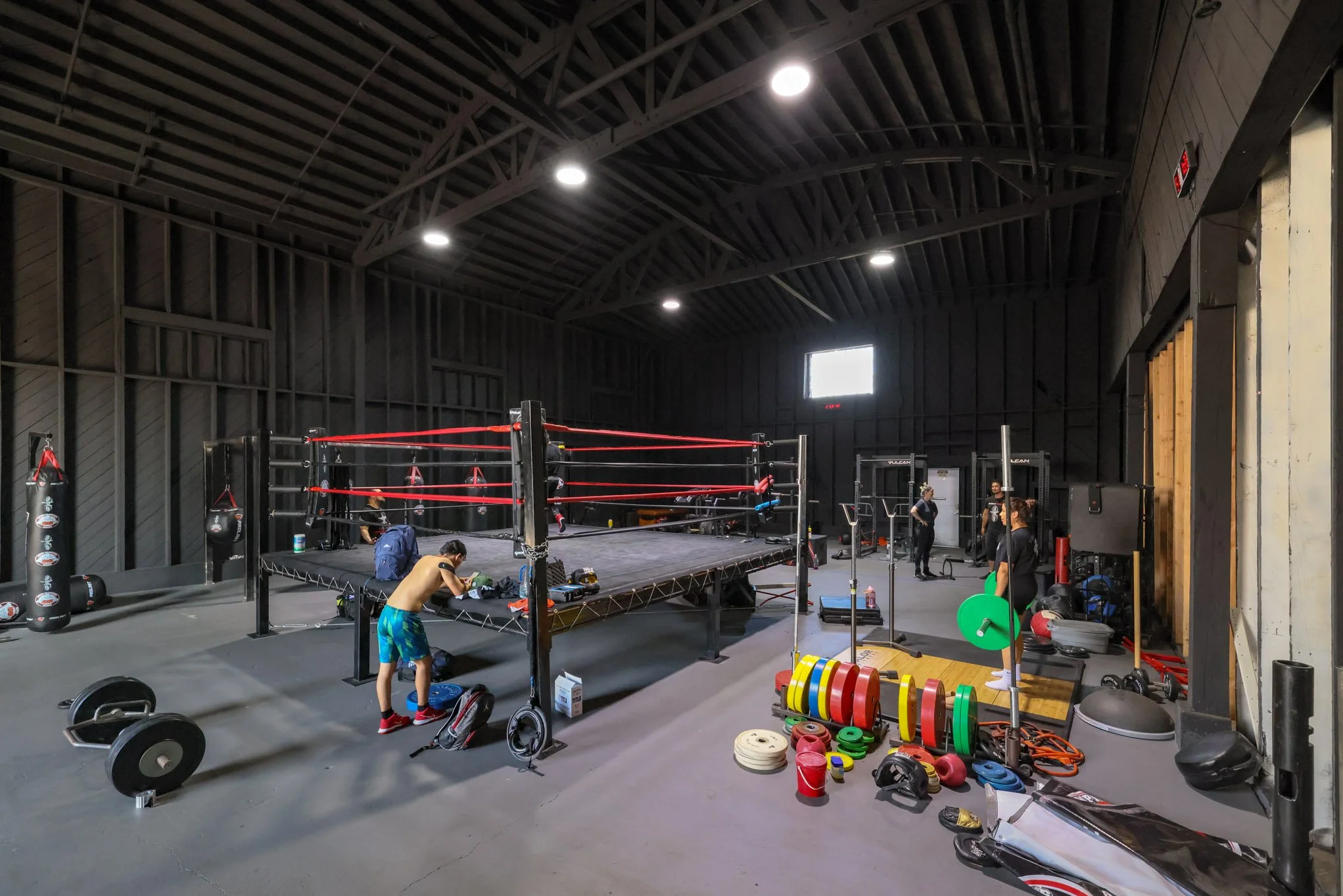 Inside a boxing gym with a boxing ring, weightlifting equipment, and a group of people training.