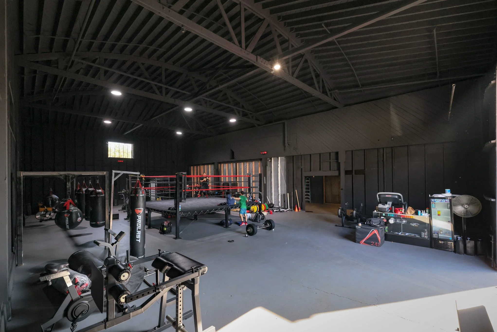 Interior of a boxing gym with a boxing ring, punching bags, workout equipment, and a few people training.
