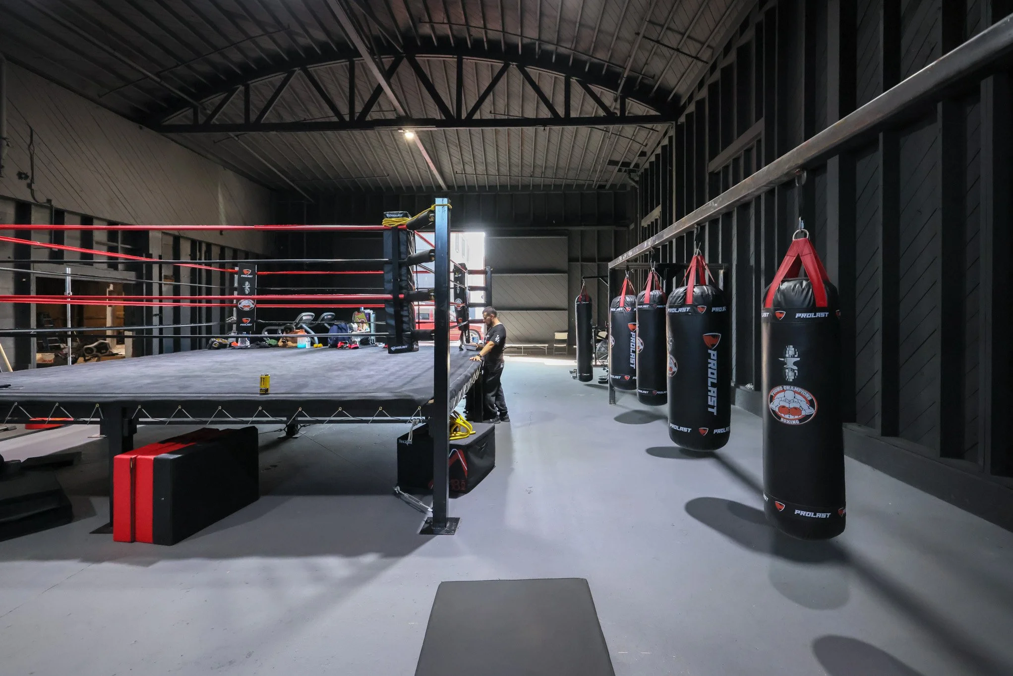 An empty boxing gym with a boxing ring in the center, punching bags hanging on the wall to the right, and equipment on the ring floor. A person is standing near the ring.