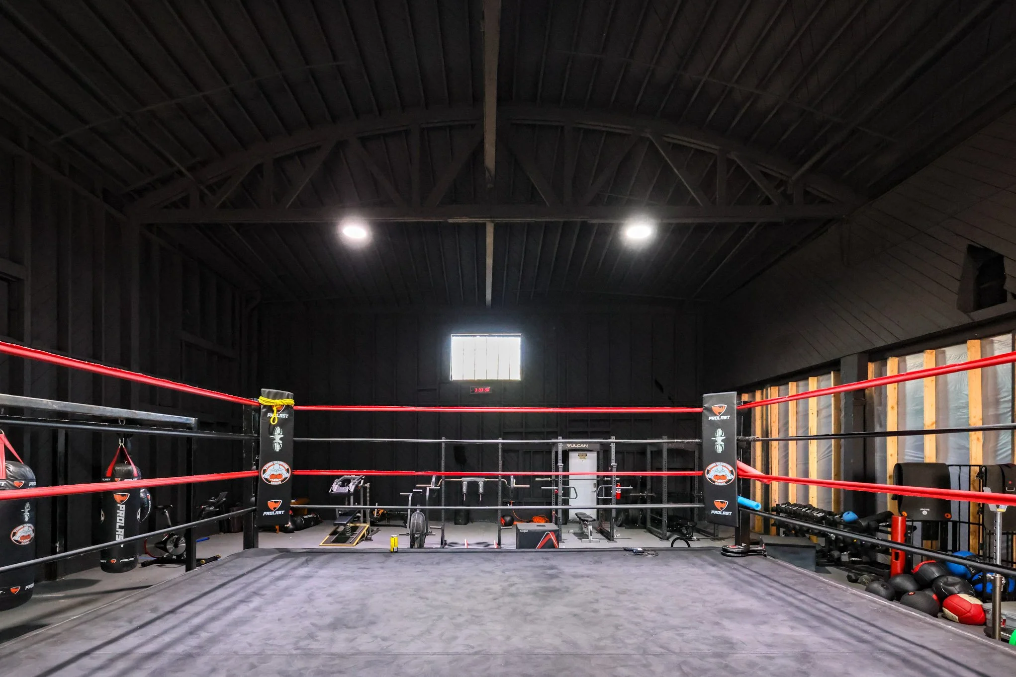 Empty boxing gym with a boxing ring, punching bags, and workout equipment in a dimly lit space.