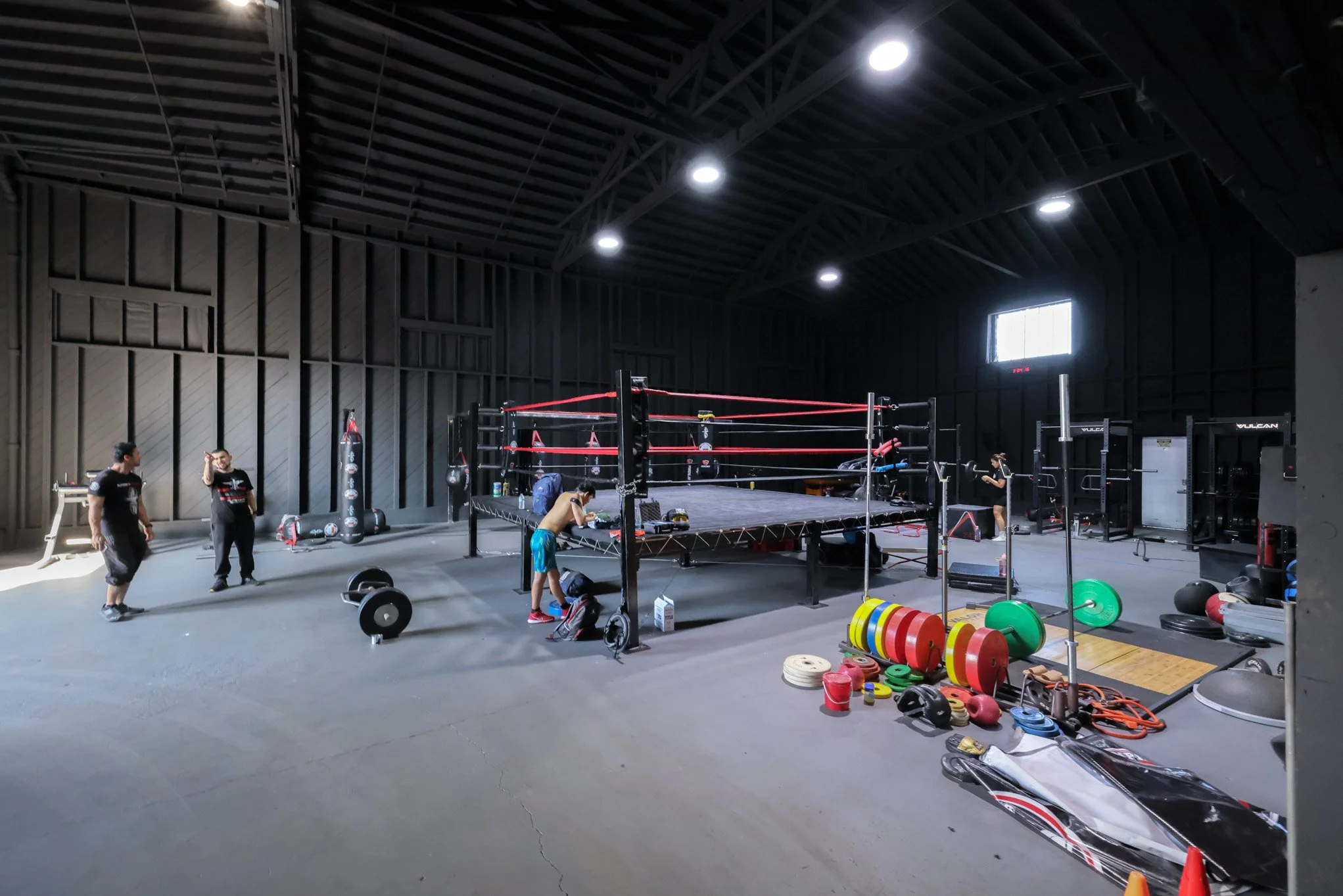 Interior of a gym with a boxing ring in the center, surrounded by various fitness equipment and a few people preparing for workout.