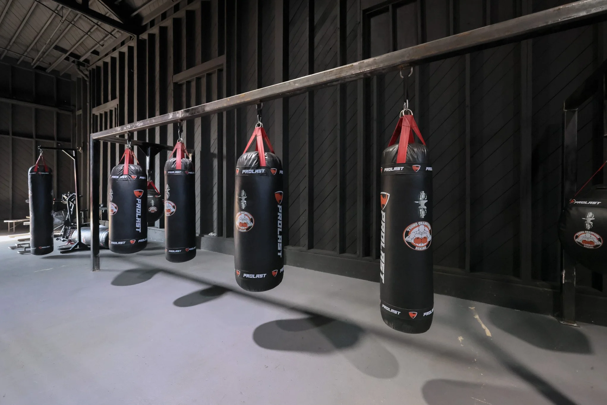 Several black punching bags hanging from a metal rail in a gym. The gym has black walls and a gray floor, with some boxing and workout equipment visible in the background.