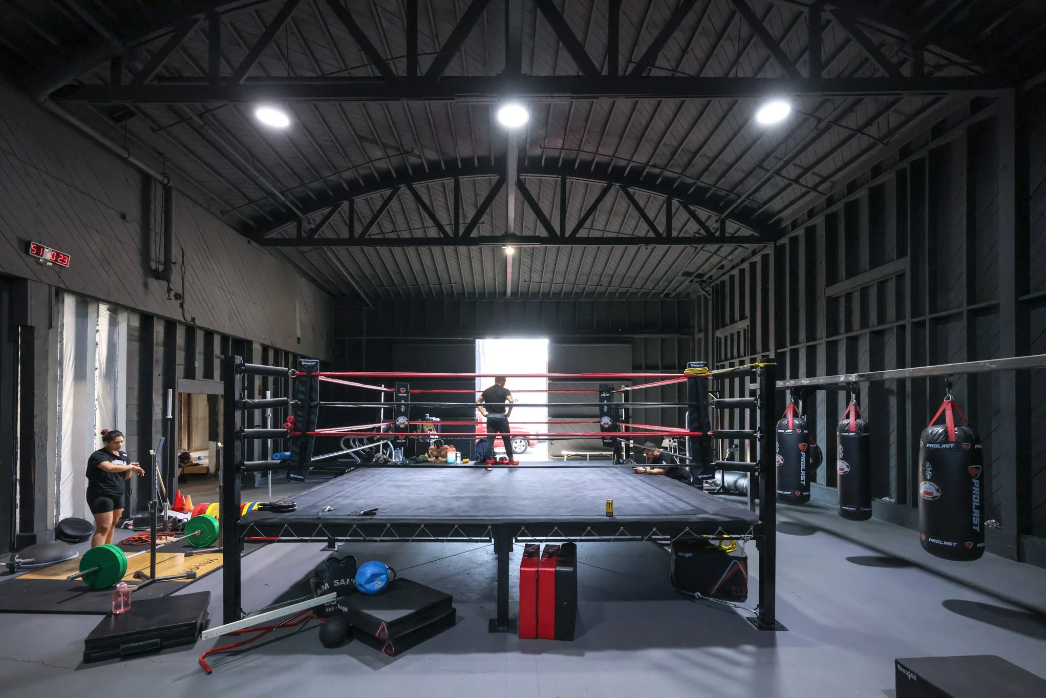 An empty boxing gym with a boxing ring in the center, hanging punching bags on the right, and people preparing equipment on the left, inside a large industrial-style building with a high metal ceiling.