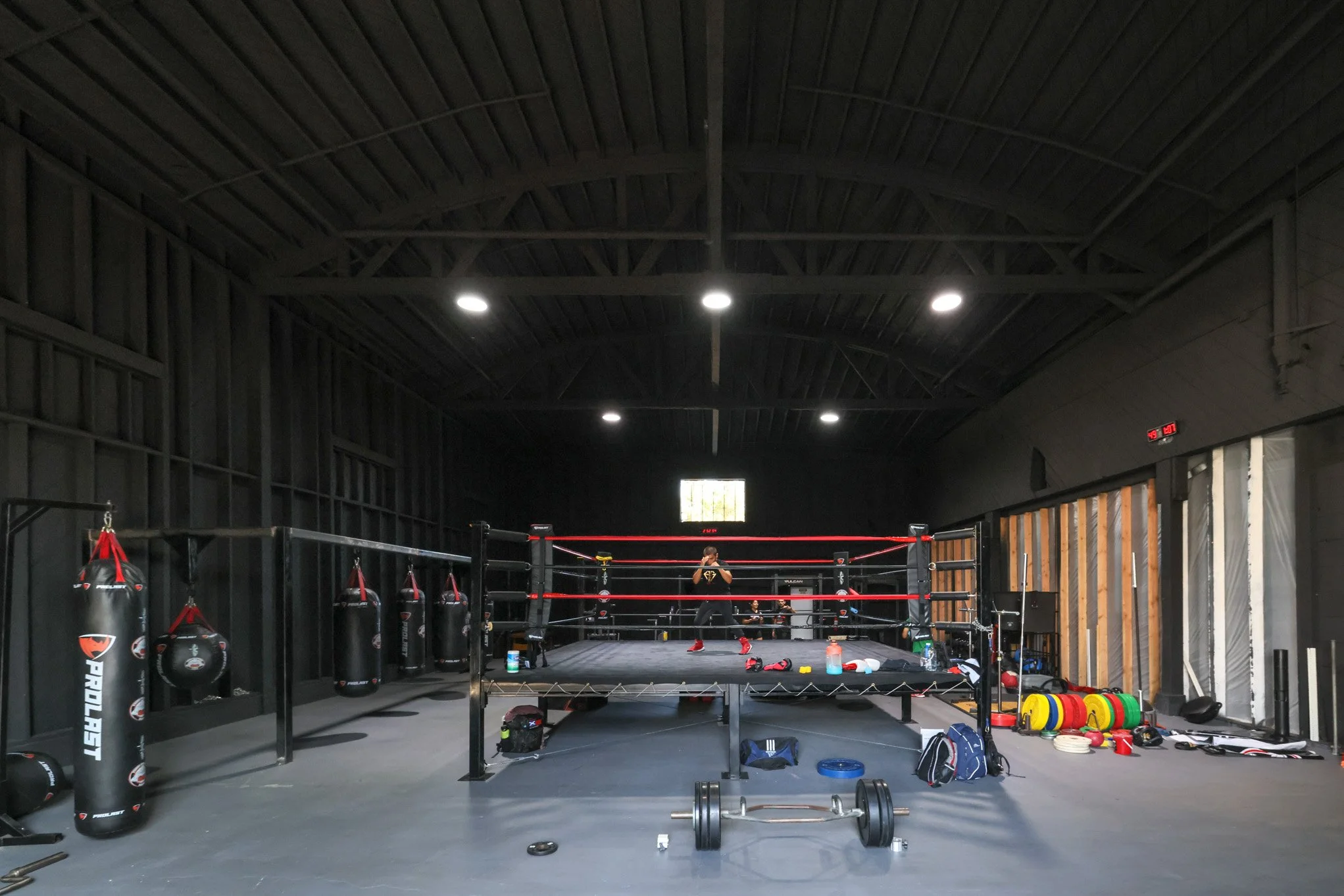 An indoor boxing gym with a boxing ring in the center, punching bags hanging on the left wall, and various gym equipment scattered around including weights and training gear.