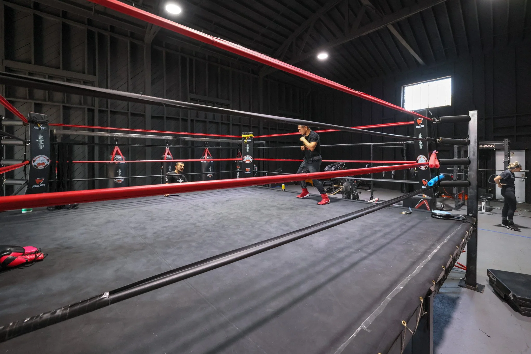 Indoor boxing or martial arts training ring with two individuals practicing, one standing in a fighting stance, and the other sitting outside the ring, with workout equipment around.