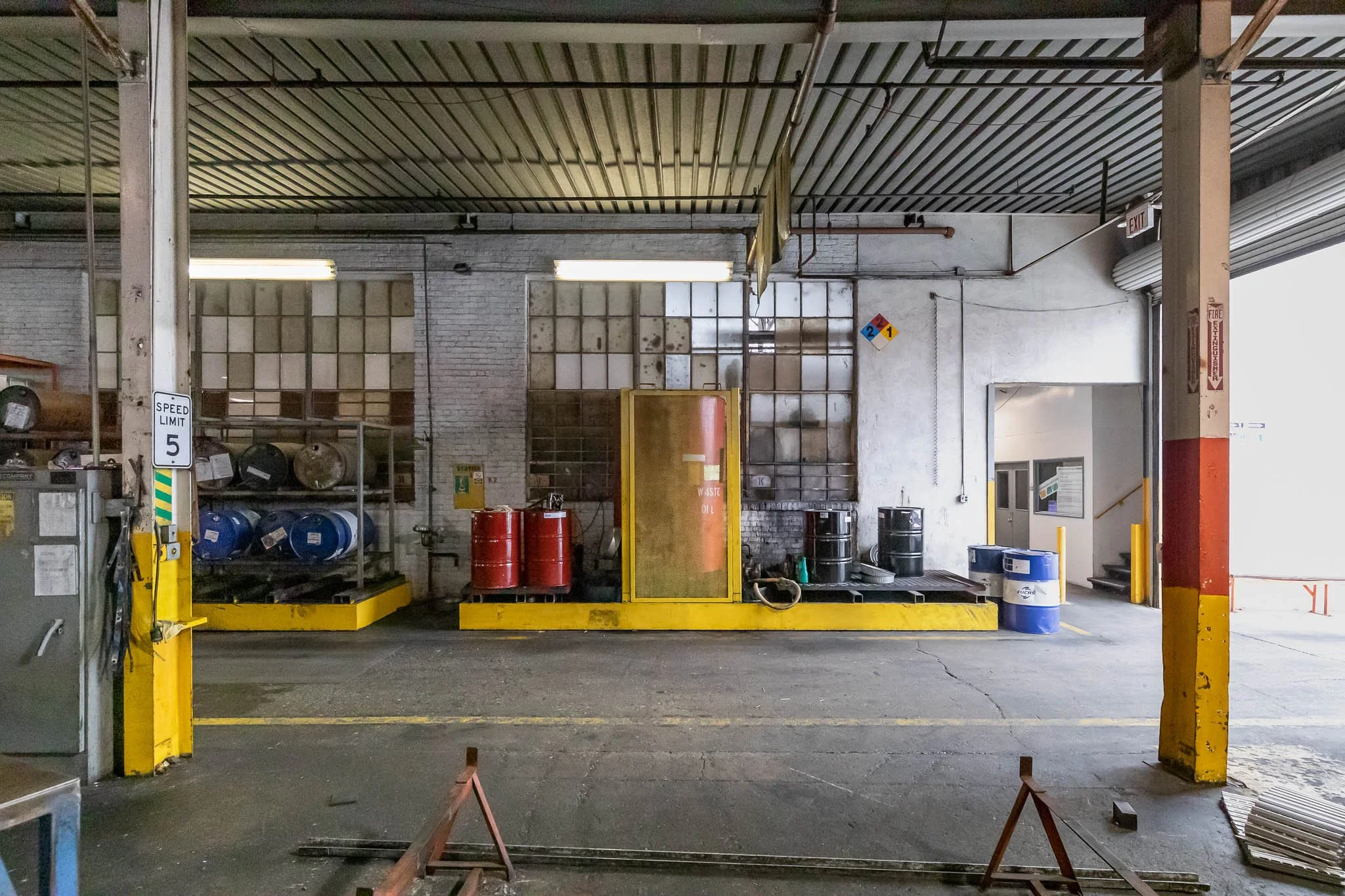 Industrial warehouse with containers, drums, and safety equipment on yellow-painted platforms, exposed brick wall behind, and a partially open rolling door on the right.