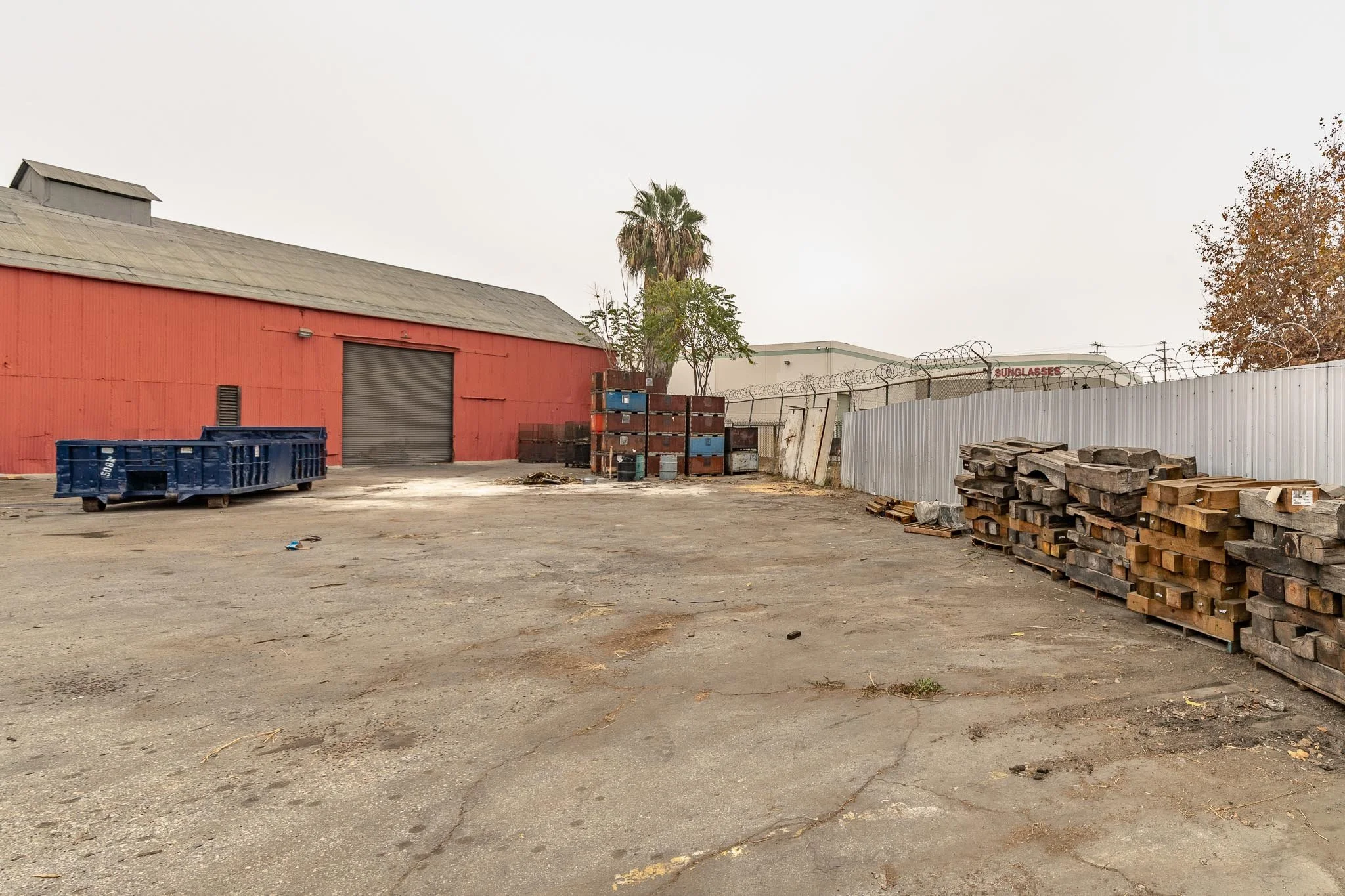 An empty industrial yard with a red warehouse, stacked wooden planks, metal containers, a blue dumpster, and a barbed wire fence.