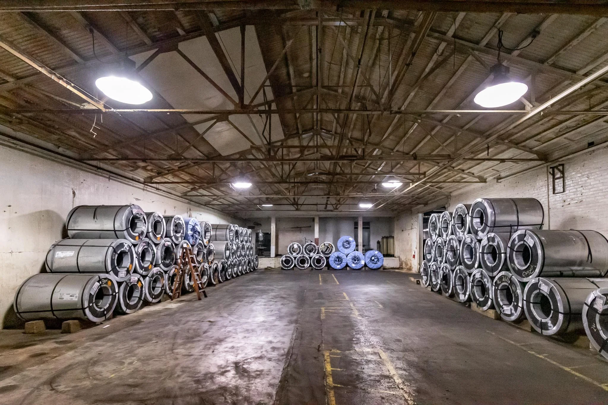 Warehouse storage with multiple metal coils stacked along the walls and on pallets, with a ladder leaning against one stack and yellow parking lines on the concrete floor.