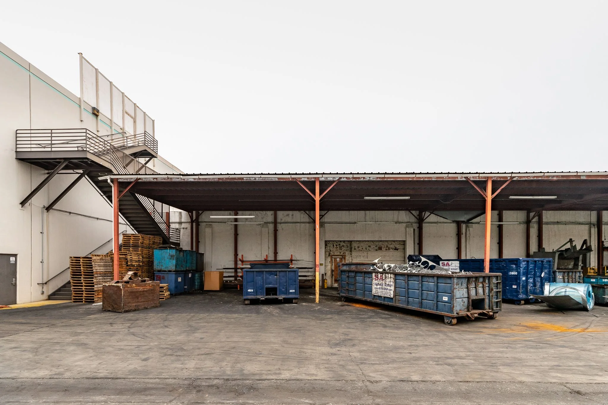 Back loading dock area with blue dumpsters filled with scrap metal, wooden pallets stacked, and a row of blue containers against a white industrial building with a metal staircase on the left side.