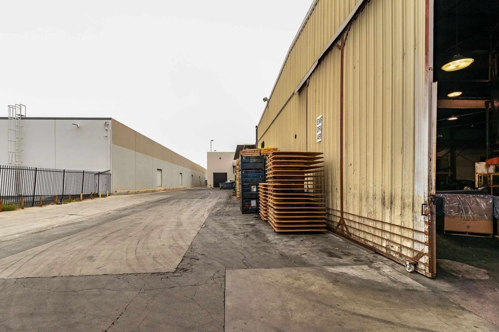 An industrial loading area with a large yellow warehouse door slightly open, revealing the inside, with stacks of wooden pallets outside and a white building in the background.