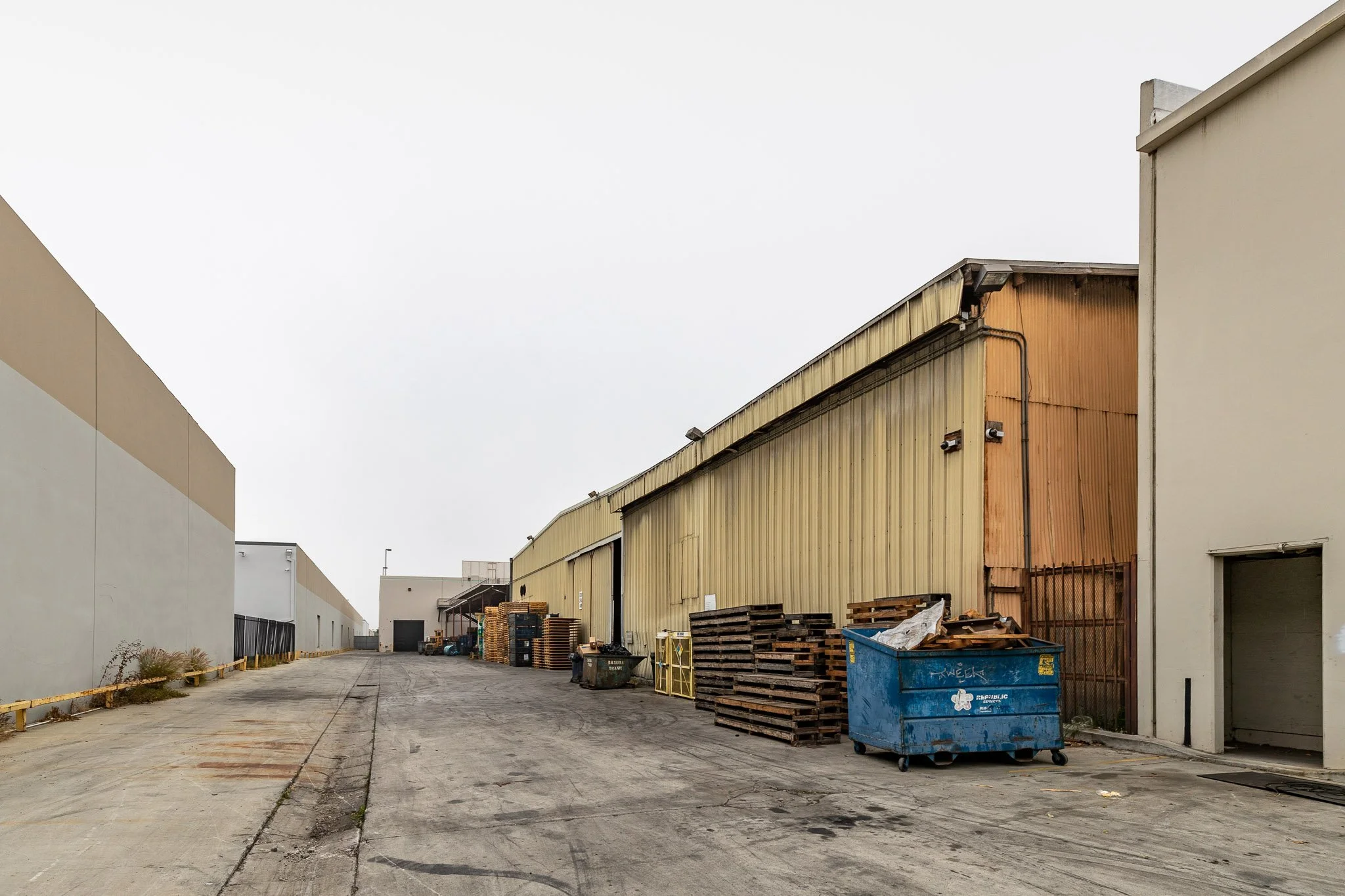 An industrial alleyway with warehouses, stacked pallets, trash bins, and scattered debris.
