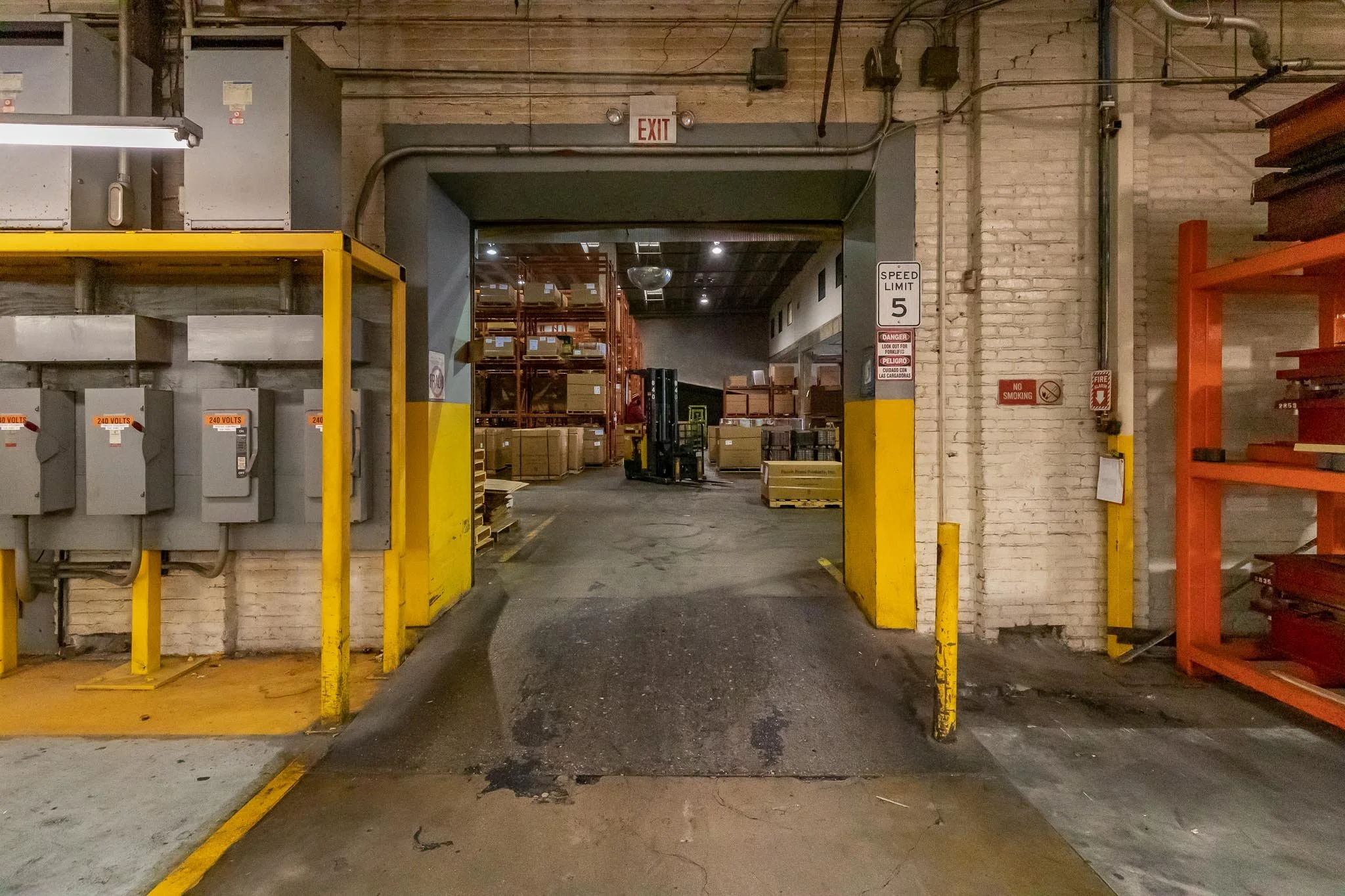 Warehouse entrance with yellow and black safety markings, electrical boxes on the left, shelving on the right, and a forklift inside, with cardboard boxes and storage racks in the background.