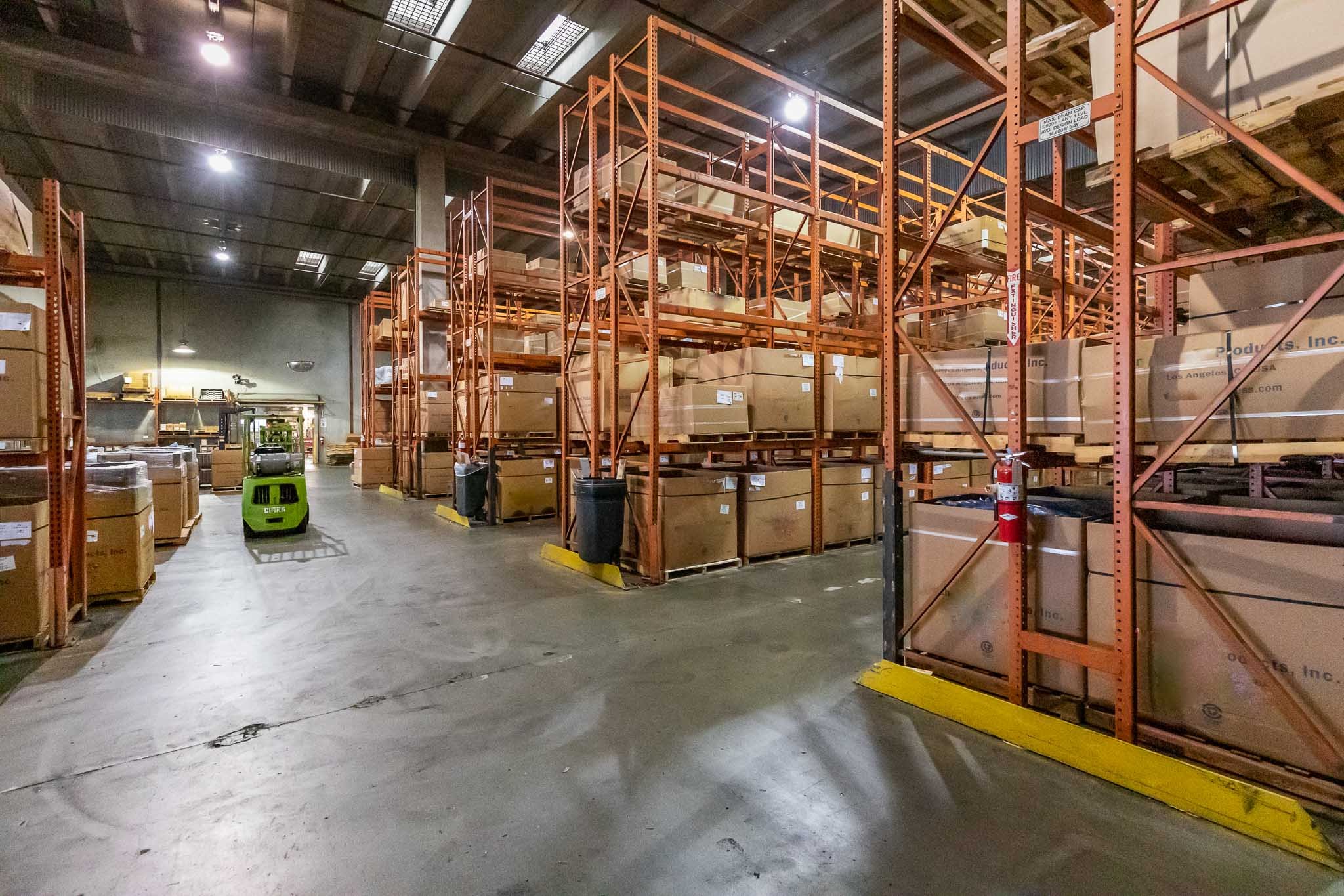 Warehouse storage area with orange metal shelving units filled with large cardboard boxes, a green forklift, and a fire extinguisher mounted on a shelf.