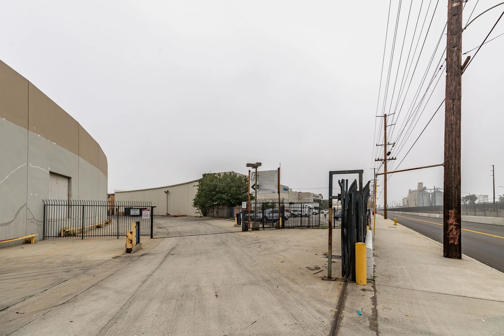Empty industrial lot with a fence, utility poles, and a road under an overcast sky.