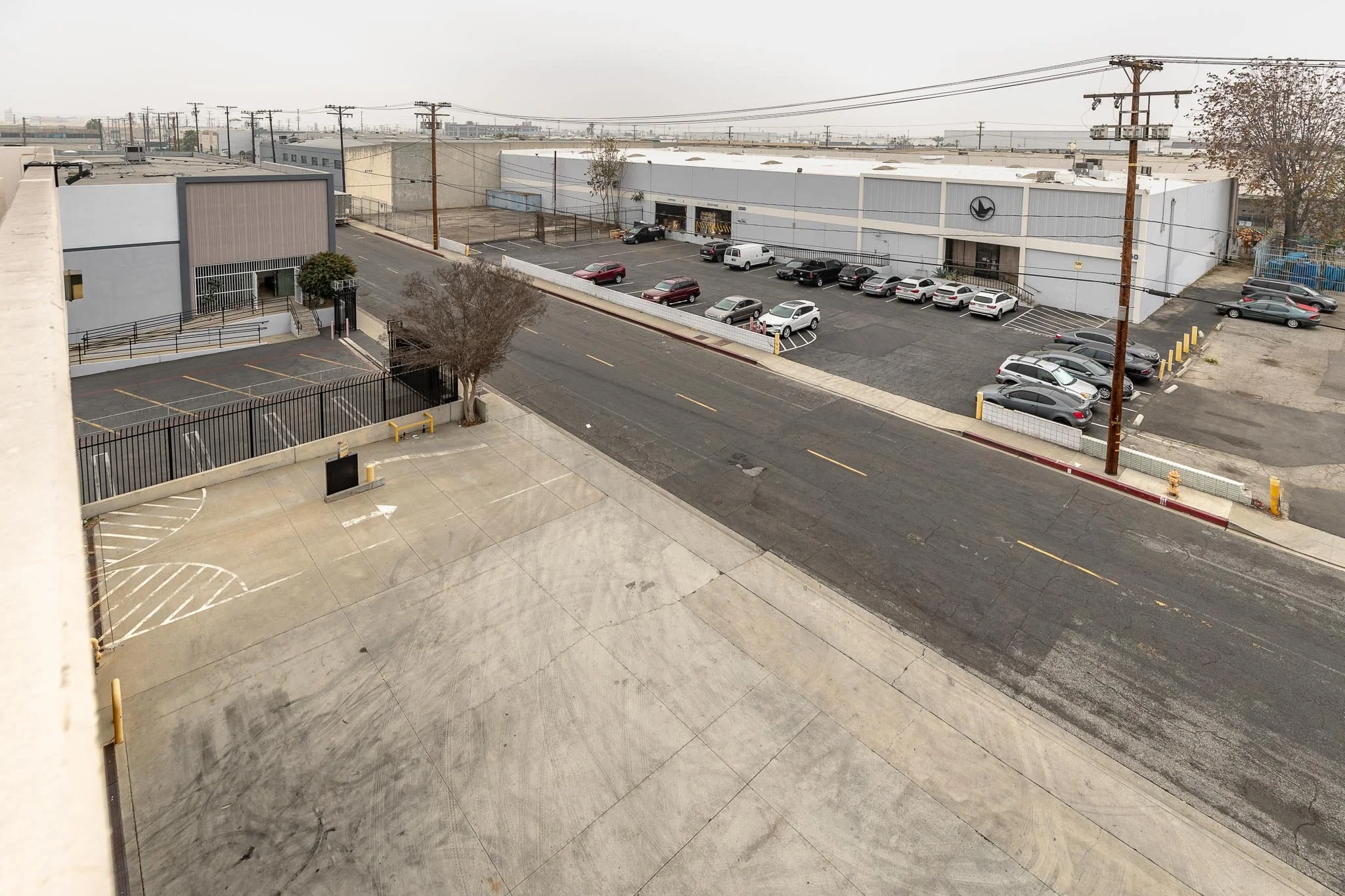 Empty parking lot with a white building on the left, a tree, and a street with parked cars and an industrial warehouse in the background.
