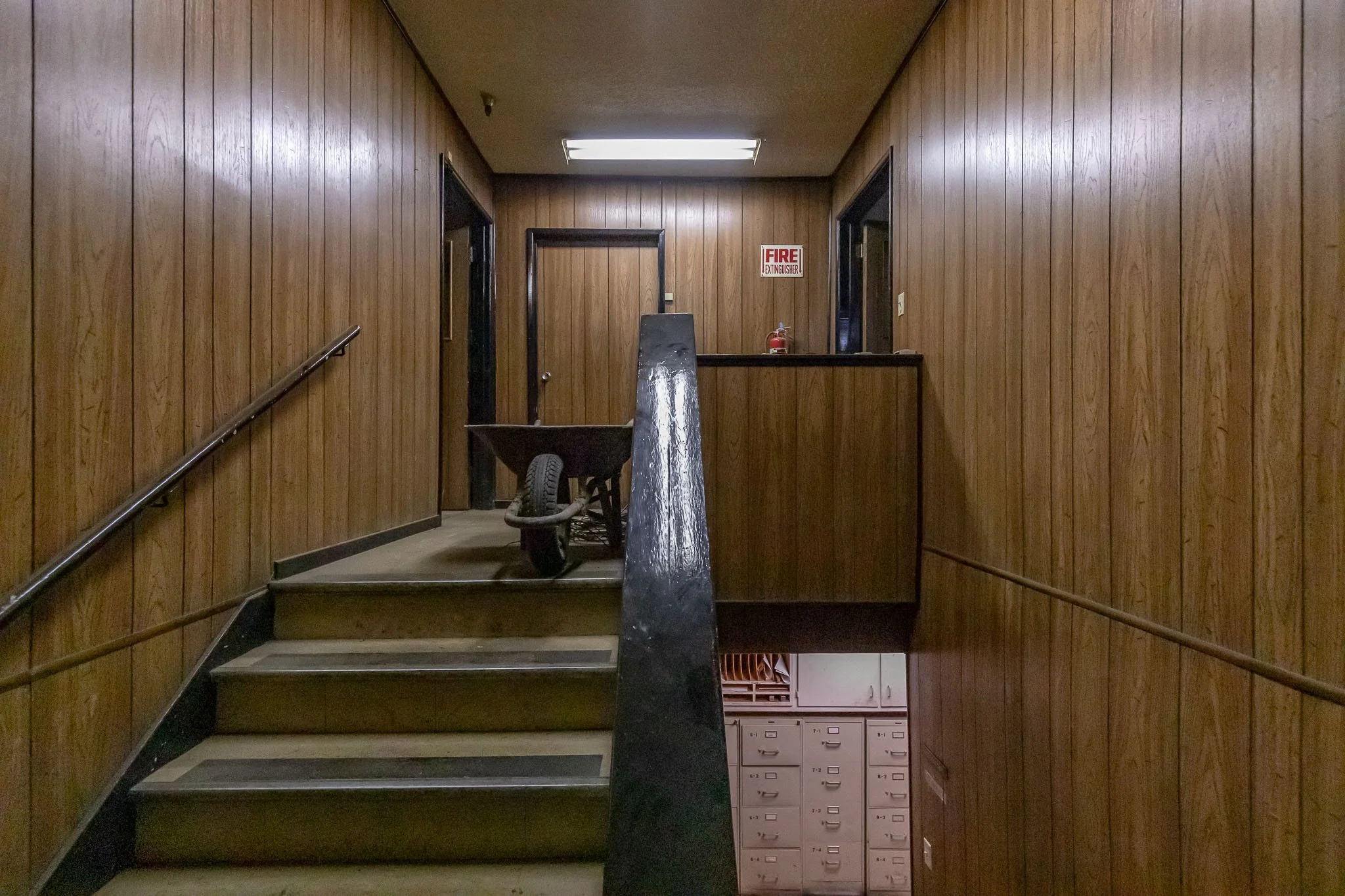 Wood-paneled interior stairwell leading to a second-floor hallway with a wheelbarrow at the top of the stairs and a fire extinguisher on the wall.