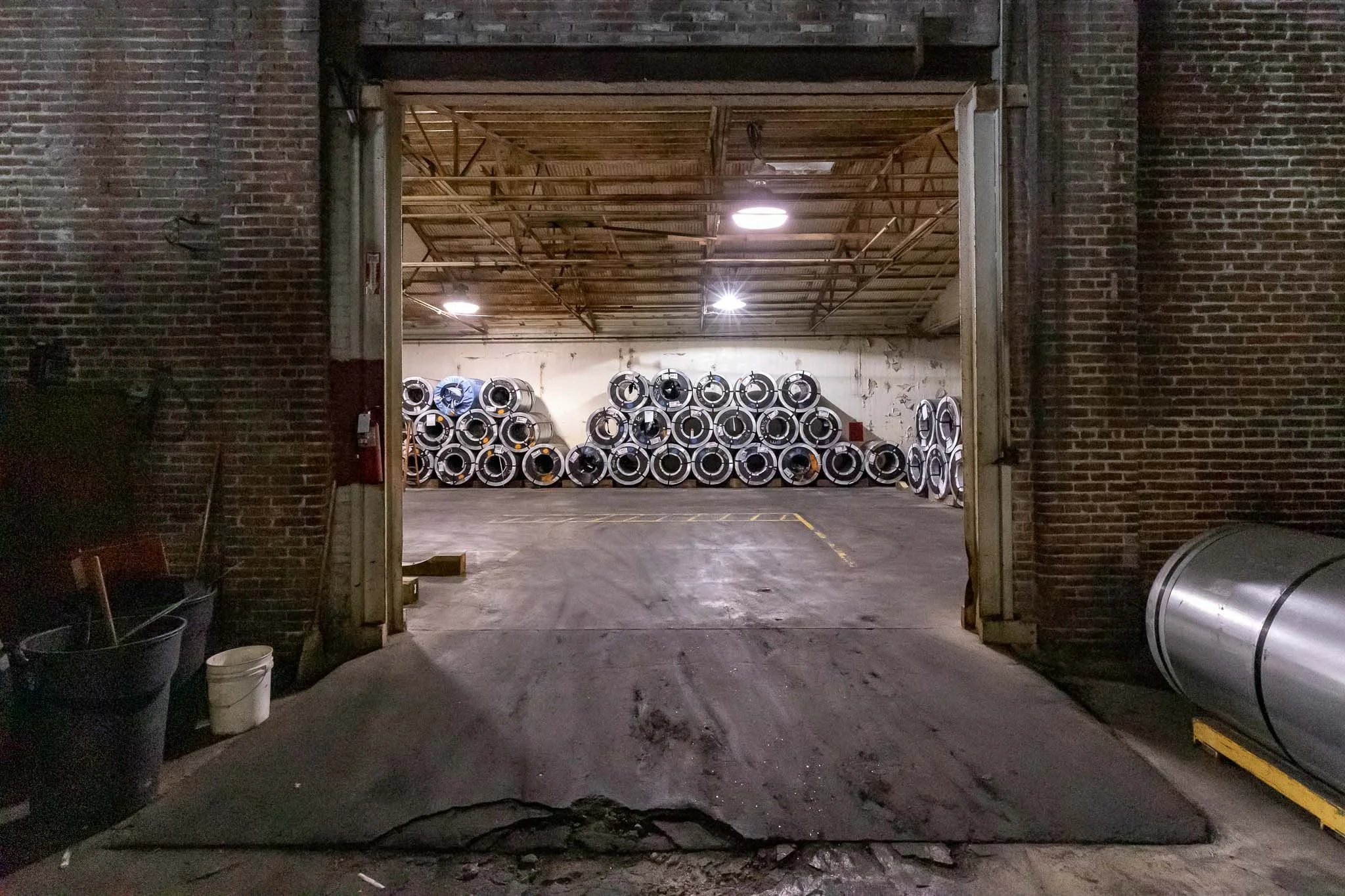 View of an industrial warehouse with stacked large metal rolls or drums against the back wall, seen through an open garage door with brick exterior.