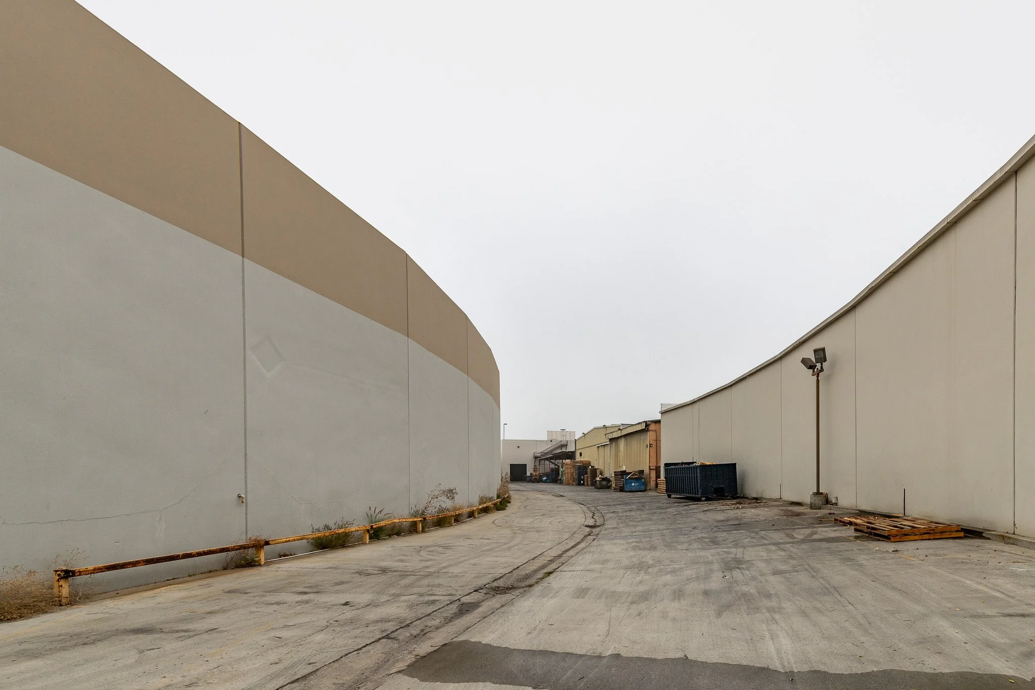 Empty alleyway between beige warehouse buildings with some trash and pallets, under an overcast sky.