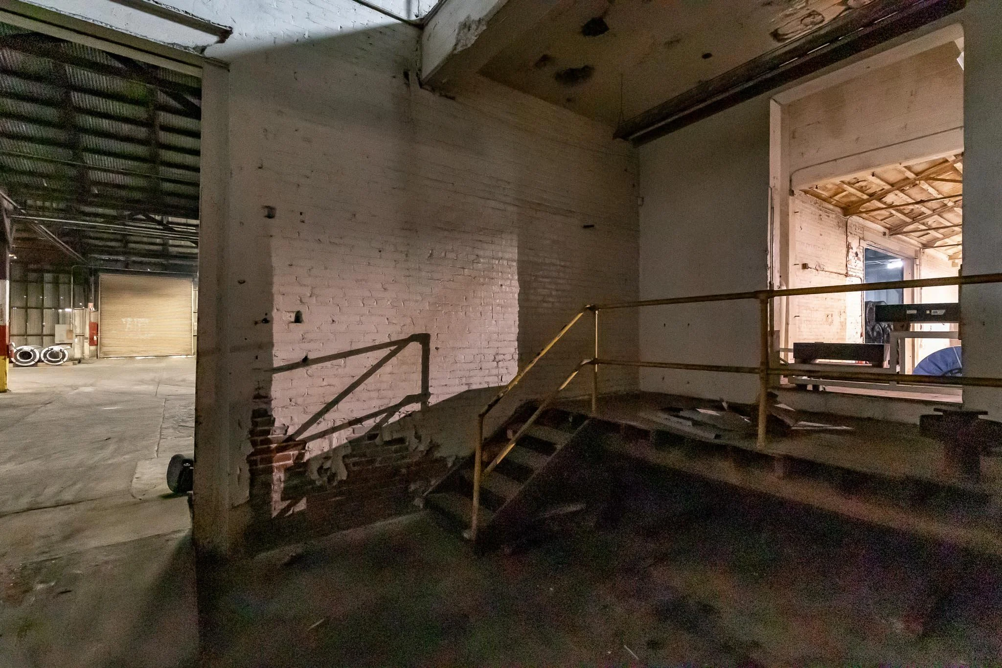 Dimly lit warehouse interior with a brick wall, metal staircase, and a view through an open doorway to another area with a ceiling featuring exposed beams.