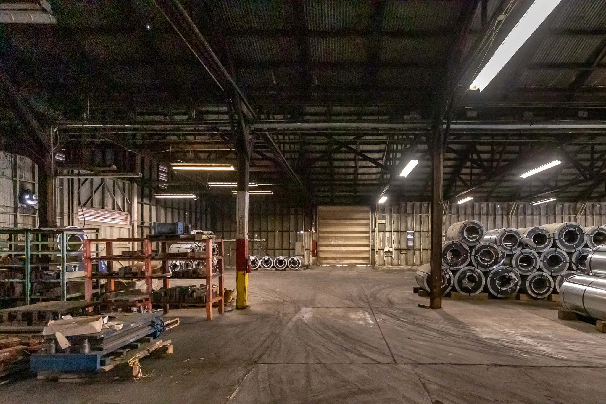 Inside a dimly lit industrial warehouse with stacks of metallic rolls and shelves.