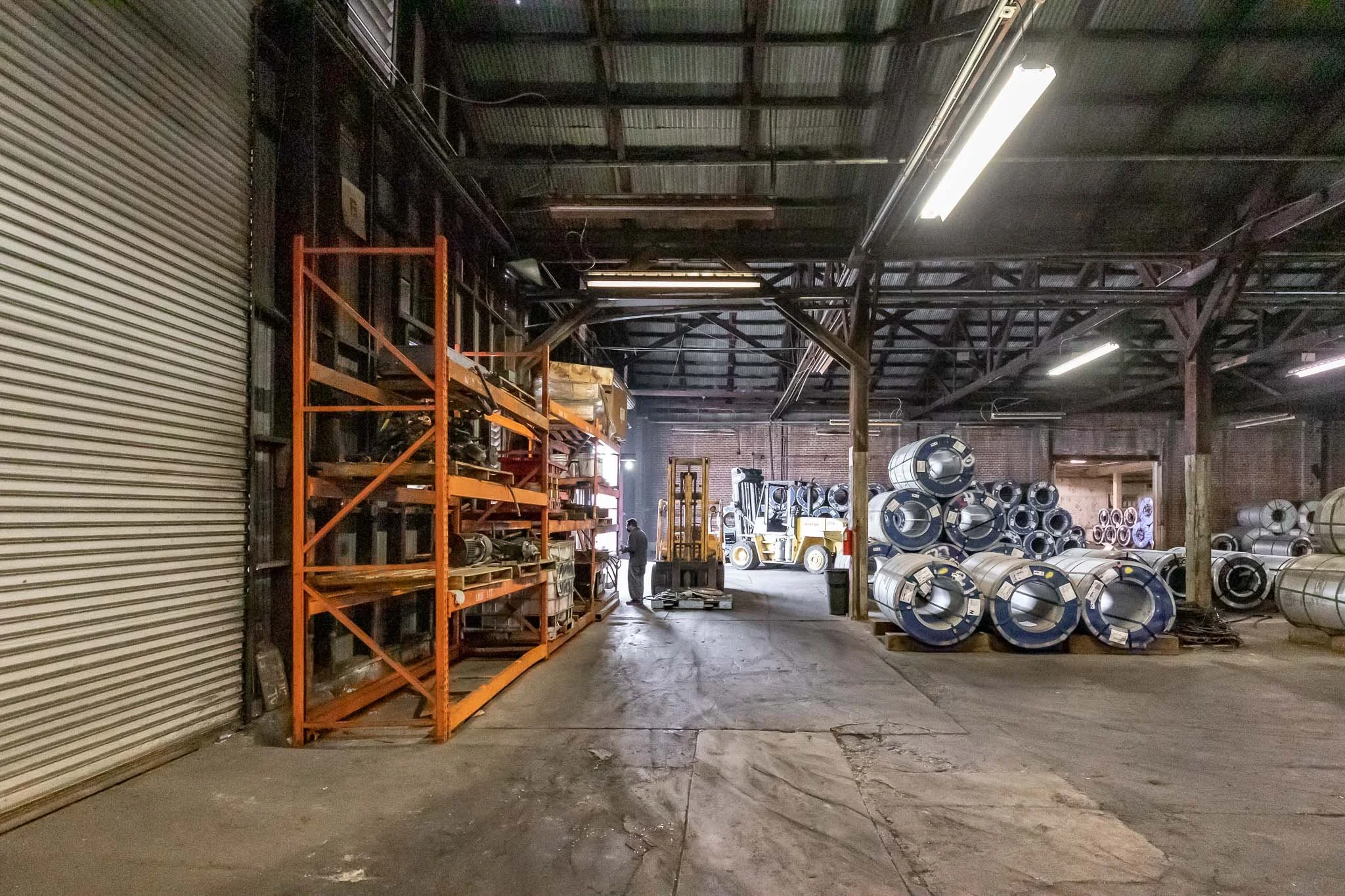 Interior of a warehouse storage area with orange shelving holding various industrial parts on the left, large metal coils stacked on pallets on the right, and a forklift near a person in the background, with a metal roll-up door on the far left.