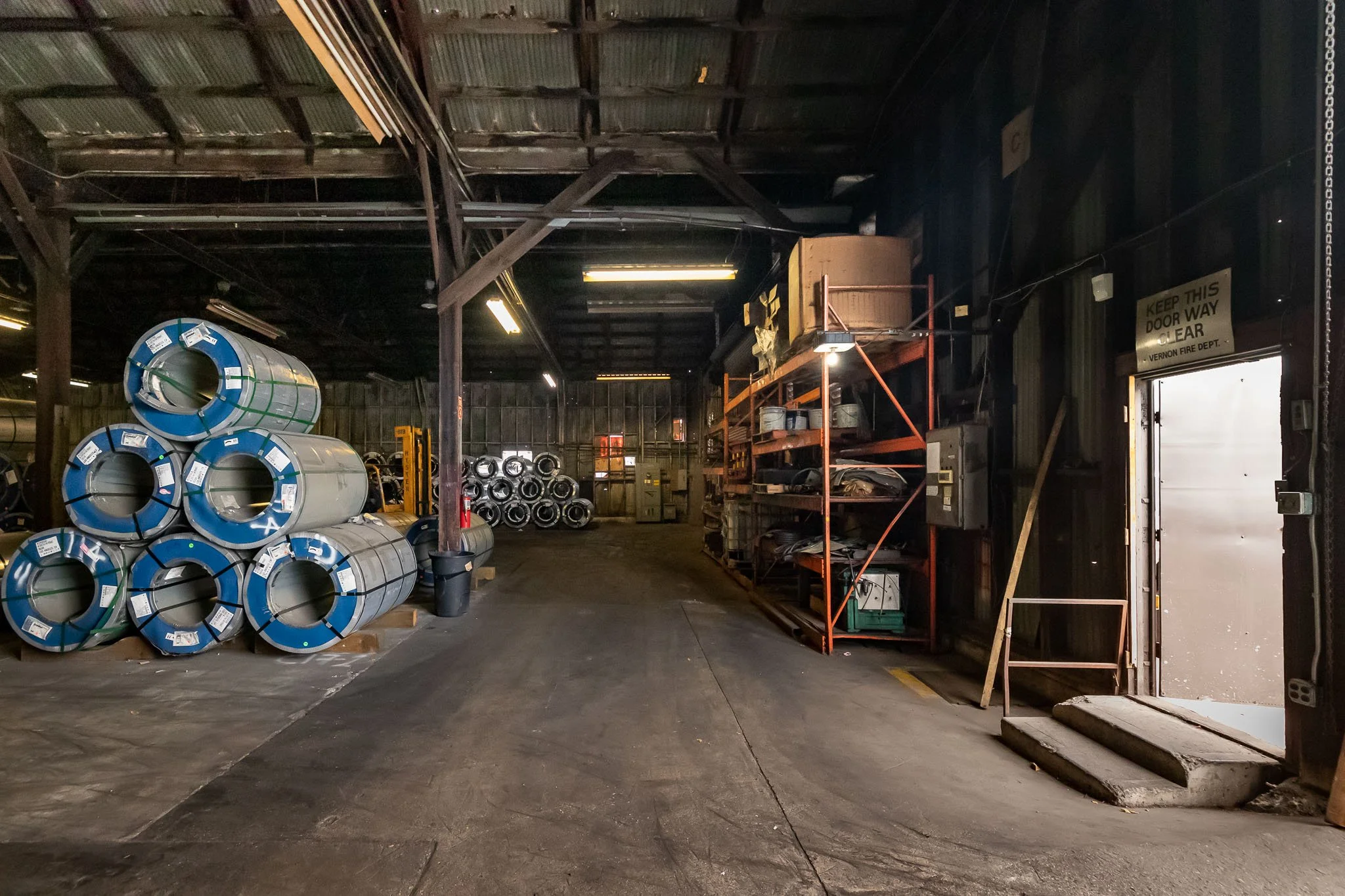 Interior of a warehouse with stacked metal pipes on the left and industrial shelving on the right, leading to a door marked 'KEEP THIS DOOR WAY CLEAR' with steps in front.