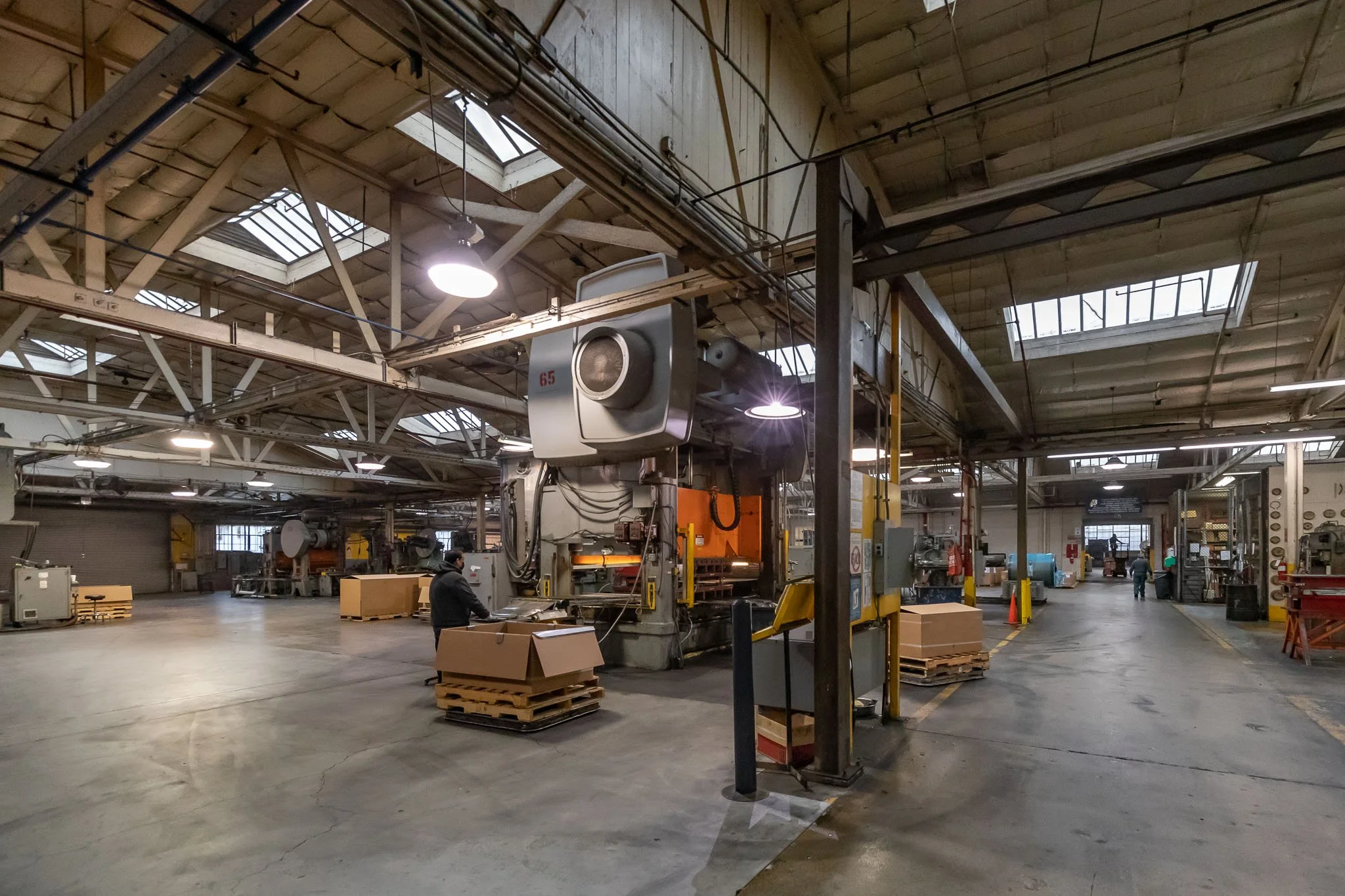 Interior of a factory or warehouse with large machinery, workers, and stacks of cardboard boxes on pallets.