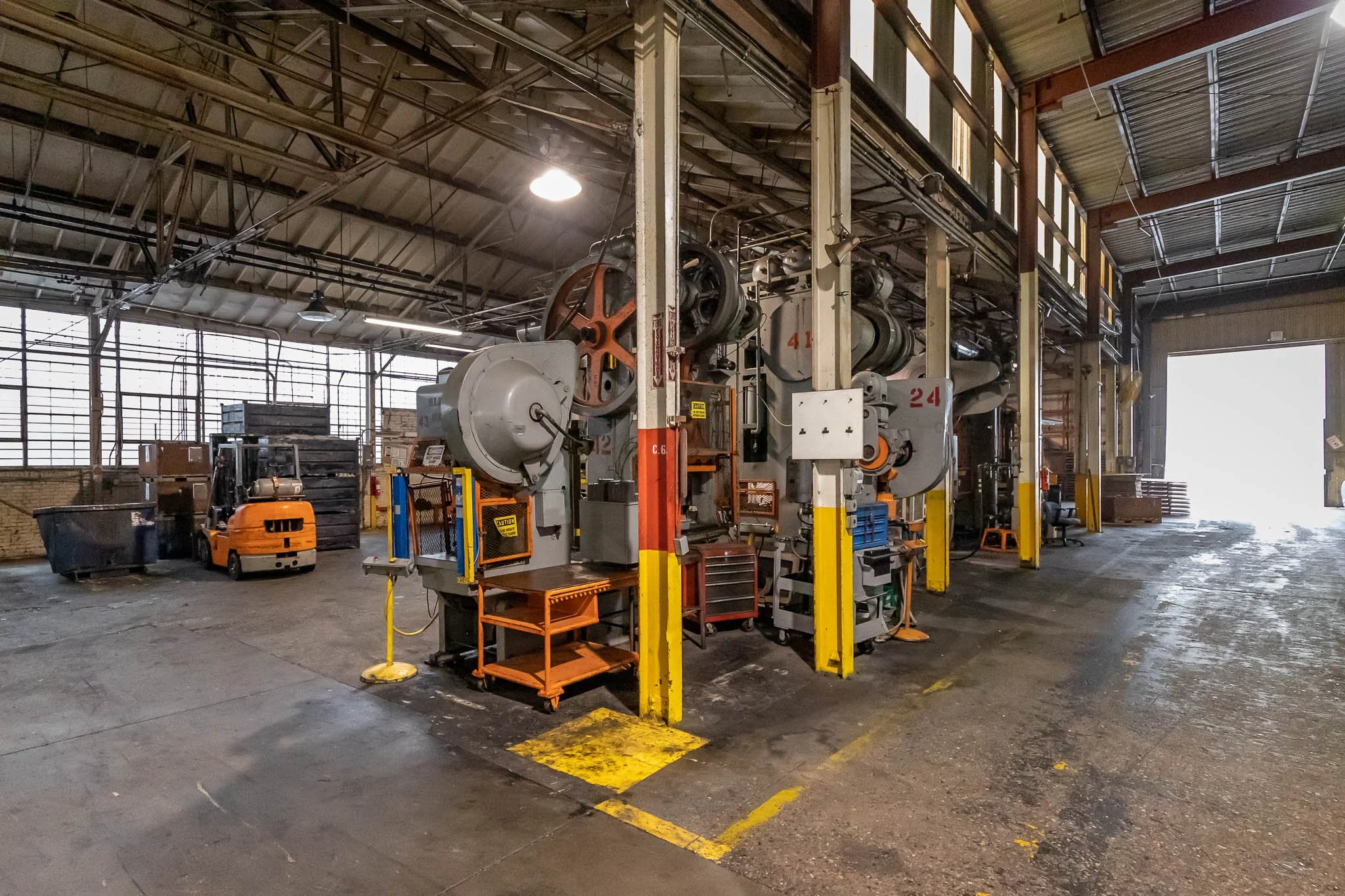 Industrial warehouse with large machinery, forklifts, pallets, and an open door on the right letting in light.