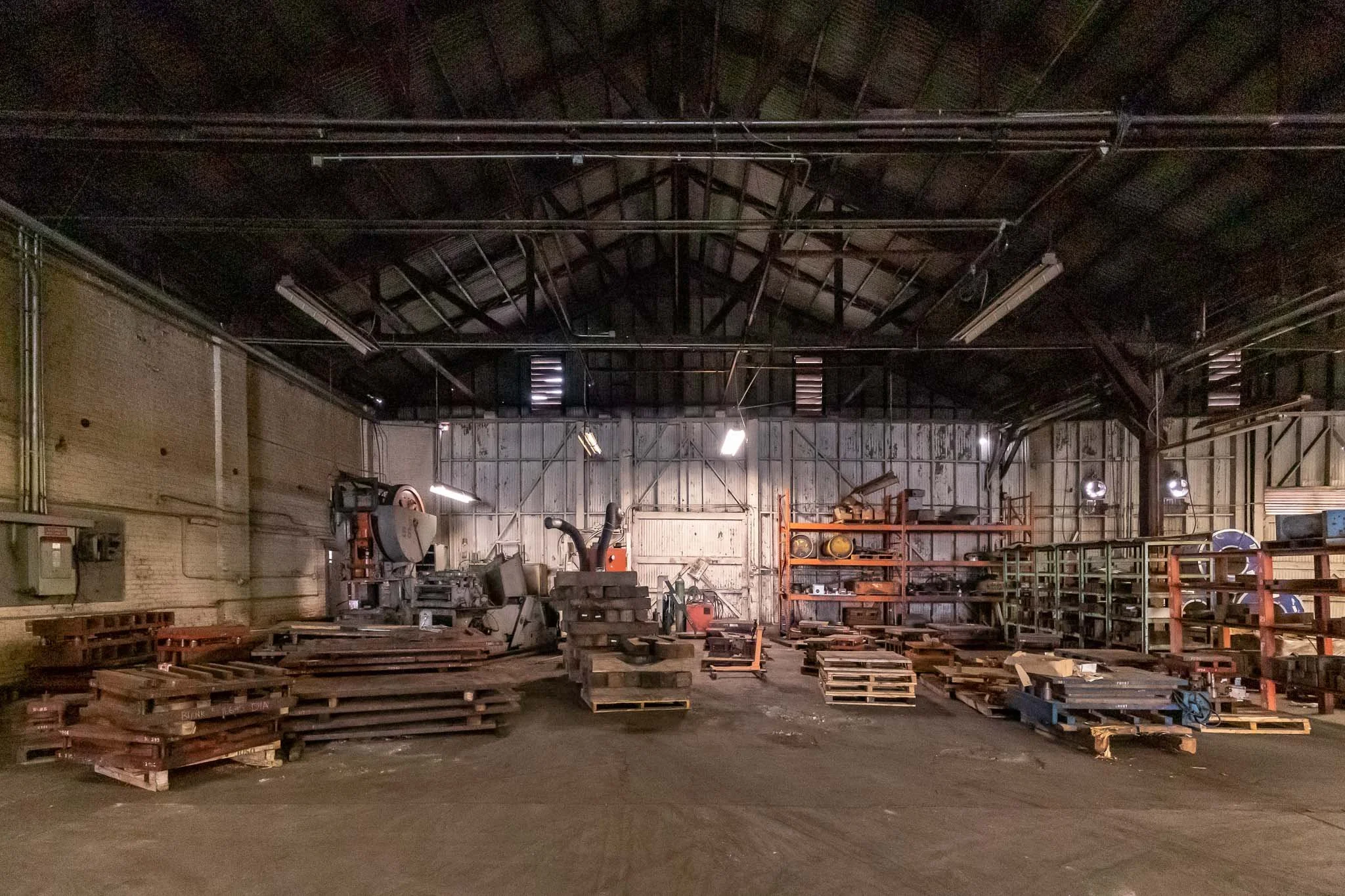 Empty industrial workshop with metal shelving, wooden pallets, and manufacturing equipment, dimly lit with fluorescent lights.