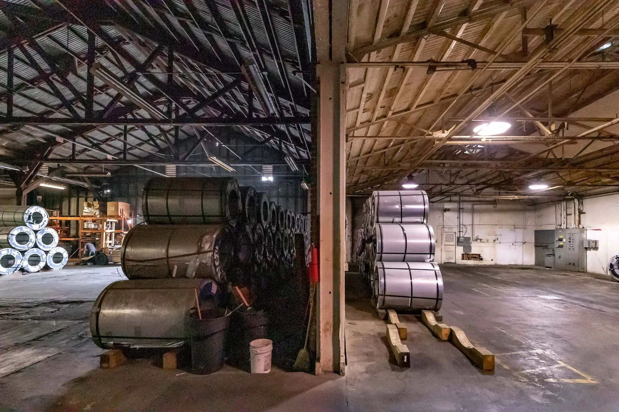 Industrial warehouse with metal rolls stacked on the floor, divided into two sections by a central wooden post, with shelves and equipment in the background.