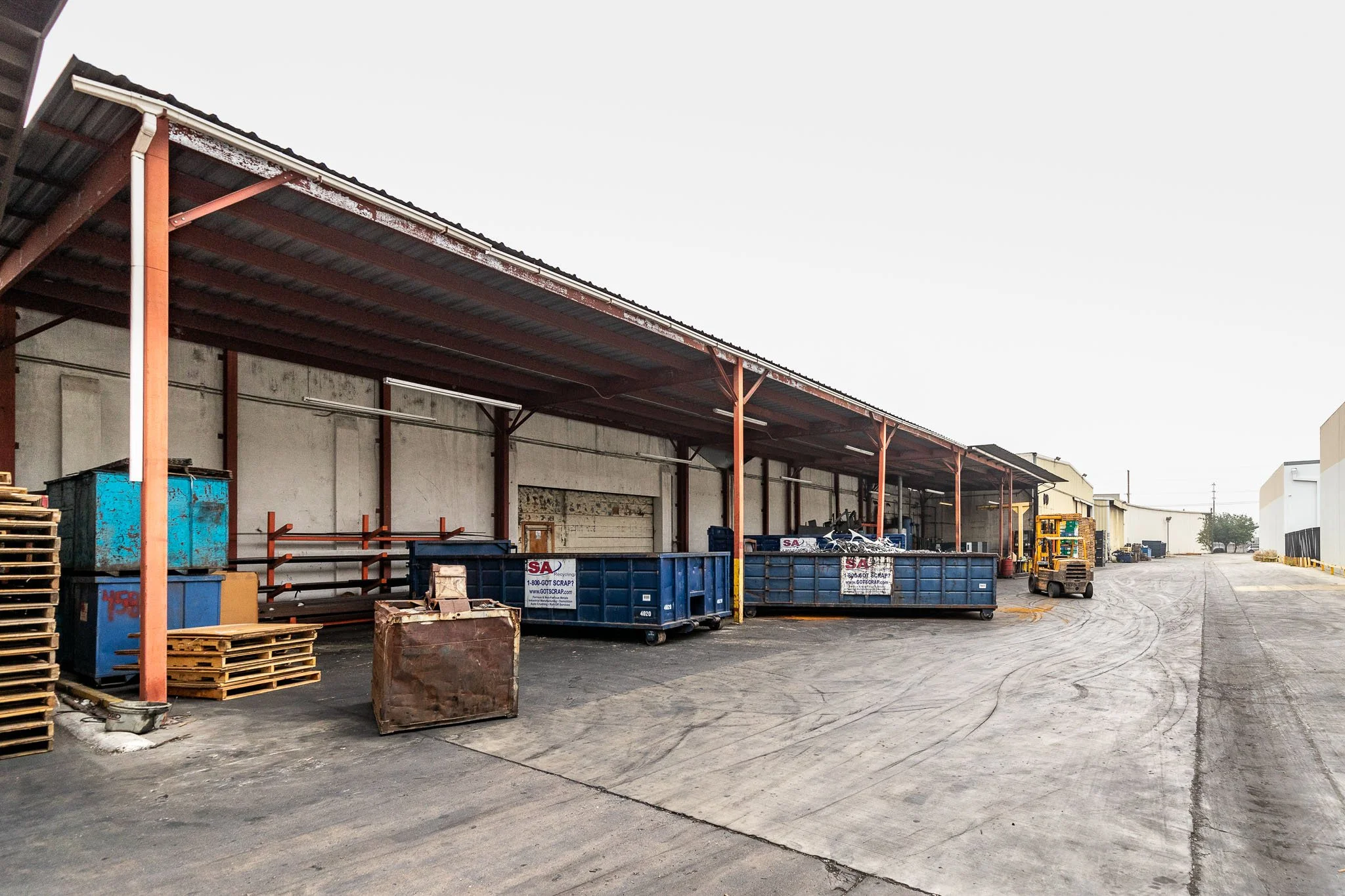 Empty industrial loading dock area with stacked pallets, large blue dumpsters, and a forklift, under a metal roof.
