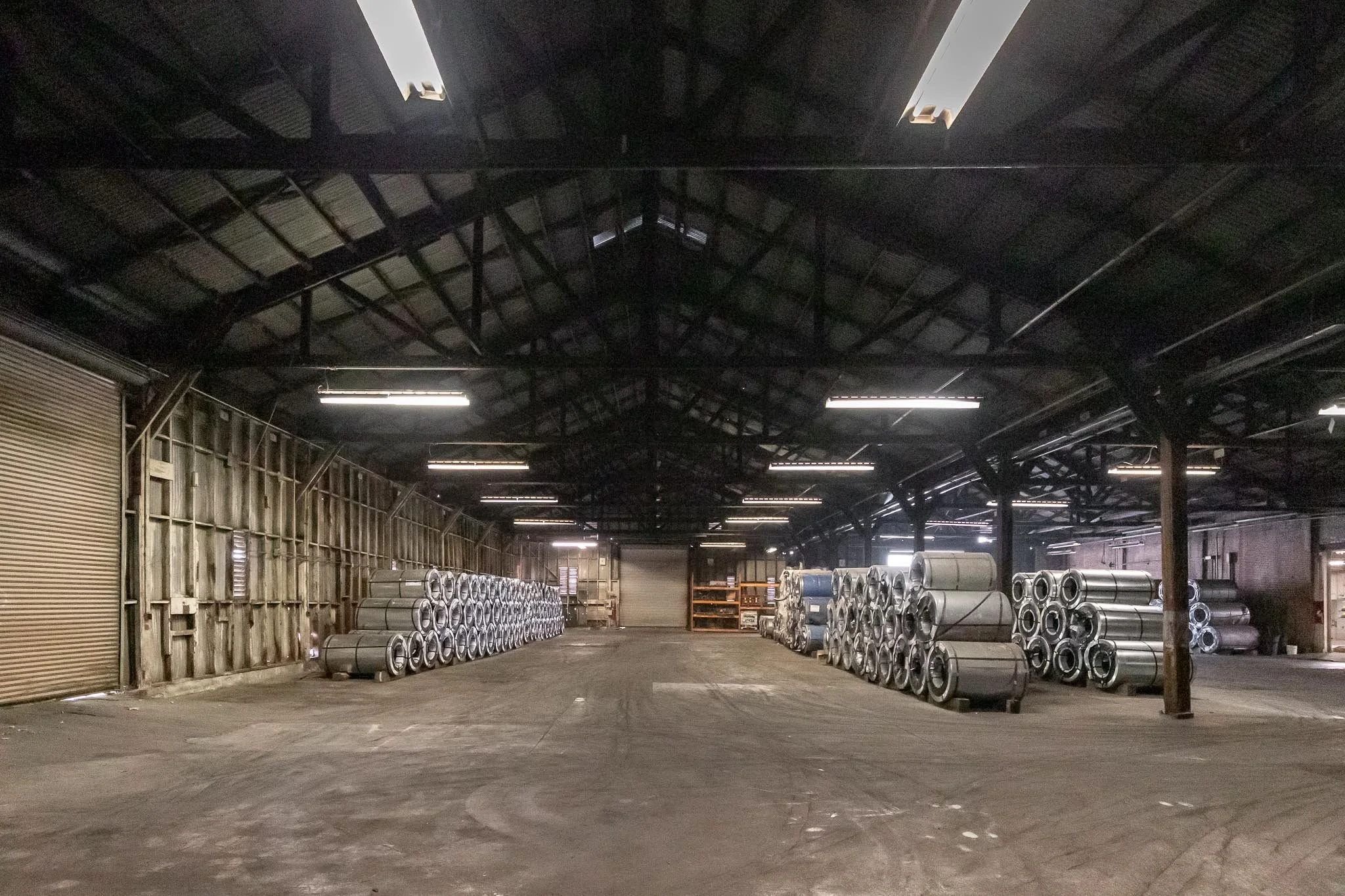 Large warehouse with stacked metal pipes along the walls, dim lighting, and a concrete floor.