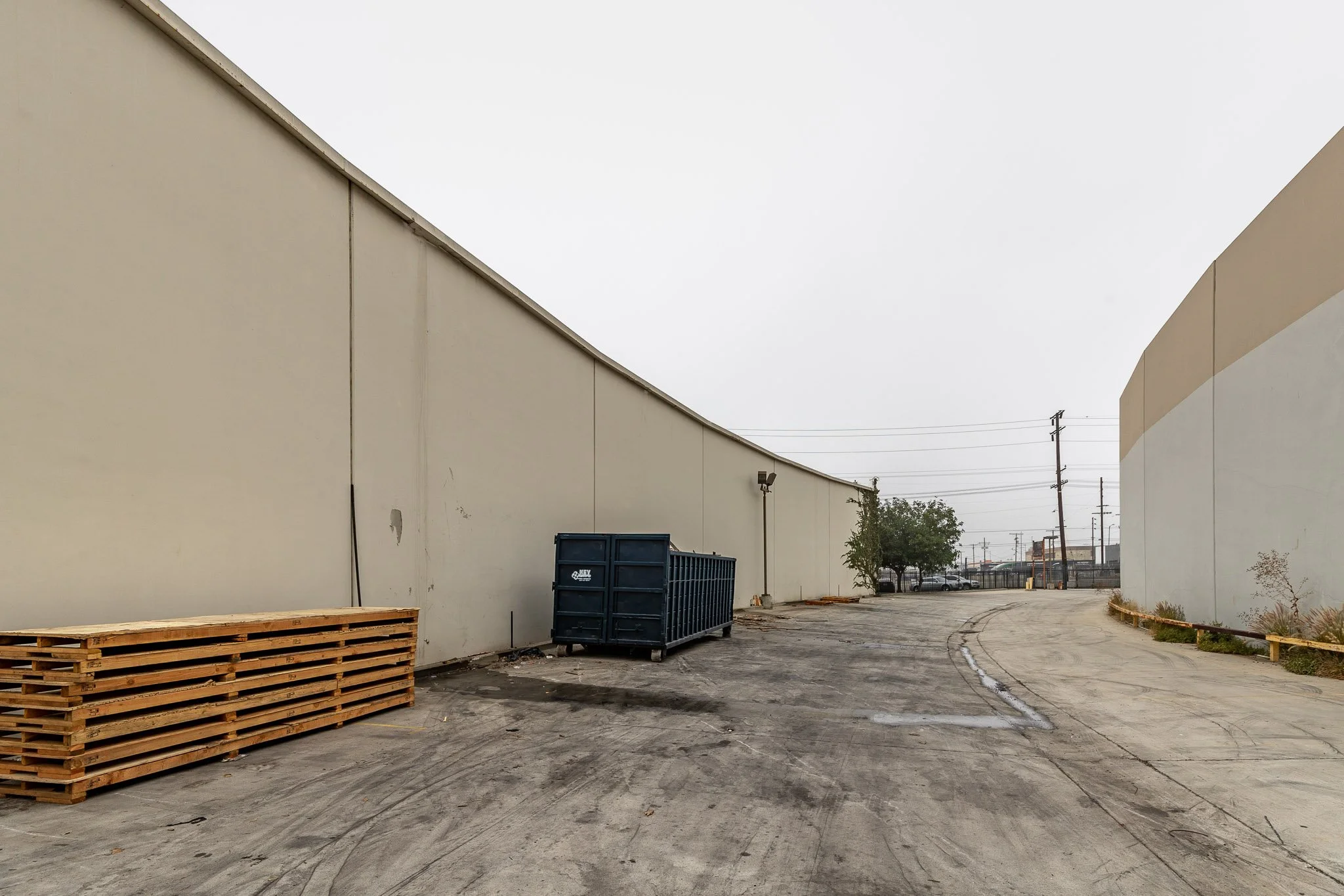 Empty industrial alleyway with tall beige walls, a large black dumpster, wooden pallets, and minimal vegetation.